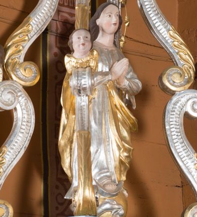 Zdjęcie nr 1: Feretron składa się z wolnostojącej, pełnoplastycznej rzeźby Matki Boskiej Niepokalanej stojącej na postumencie oraz fazowanym i profilowanym cokole, ponad którą wznosi się baldachim wsparty na czterech, spiętrzonych esowato wygiętych i srebrzonych zdwojonych wolutach. Woluty zdobią stylizowane, złocone palmety oraz rzeźbione, uskrzydlone główki aniołków, umieszczone na ich łączeniu, a także przy płaskim baldachimie, na którym spoczywa drewniana poduszka z koroną zamknietą. Całopostaciowa figura Marii ujęta została frontalnie w lekkim kontrapoście z modlitewnie złożonymi rękami uniesionymi na wysokości piersi. Niepokalana prawą stopę wspiera o srebrny  półksiężyc leżący na złotym globie, opleciony srebrnym wężem, który w paszczy trzyma jabłko. Jej głowa o owalnej twarzy, długiej szyi i jasnej karnacji oraz rumianych policzkach, drobnym nosie i wydatnych ustach jest nieznacznie odchylona w prawo. Długie, brązowe włosy opadają na jej plecy, odsłaniając uszy. Wokół głowy znajduje się druciany nimb podtrzymujący siedem złoconych gwiazd. Podniebie baldachimu ozdobiono płaskorzeźbionym, srebrzonym i złoconym wyobrażeniem gołębicy Ducha Świętego na tle promieni. Twarze uskrzydlonych aniołków na podporach baldachimu charakteryzują się jasną karnacją, wysokimi czołami, okrągłymi i pulchnymi policzkami oraz wydatnymi ustami. Ich głowy otaczają krótkie, brązowe włosy. Na szczycie baldachimu ustawiono rzeźbioną i srebrzoną poduszkę dekorowaną złocistym galonem i chwostami na rogach. Spoczywa na niej drewniana i złocona korona zamknięta.  