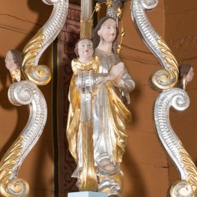 Zdjęcie nr 1: Feretron składa się z wolnostojącej, pełnoplastycznej rzeźby Matki Boskiej Niepokalanej stojącej na postumencie oraz fazowanym i profilowanym cokole, ponad którą wznosi się baldachim wsparty na czterech, spiętrzonych esowato wygiętych i srebrzonych zdwojonych wolutach. Woluty zdobią stylizowane, złocone palmety oraz rzeźbione, uskrzydlone główki aniołków, umieszczone na ich łączeniu, a także przy płaskim baldachimie, na którym spoczywa drewniana poduszka z koroną zamknietą. Całopostaciowa figura Marii ujęta została frontalnie w lekkim kontrapoście z modlitewnie złożonymi rękami uniesionymi na wysokości piersi. Niepokalana prawą stopę wspiera o srebrny  półksiężyc leżący na złotym globie, opleciony srebrnym wężem, który w paszczy trzyma jabłko. Jej głowa o owalnej twarzy, długiej szyi i jasnej karnacji oraz rumianych policzkach, drobnym nosie i wydatnych ustach jest nieznacznie odchylona w prawo. Długie, brązowe włosy opadają na jej plecy, odsłaniając uszy. Wokół głowy znajduje się druciany nimb podtrzymujący siedem złoconych gwiazd. Podniebie baldachimu ozdobiono płaskorzeźbionym, srebrzonym i złoconym wyobrażeniem gołębicy Ducha Świętego na tle promieni. Twarze uskrzydlonych aniołków na podporach baldachimu charakteryzują się jasną karnacją, wysokimi czołami, okrągłymi i pulchnymi policzkami oraz wydatnymi ustami. Ich głowy otaczają krótkie, brązowe włosy. Na szczycie baldachimu ustawiono rzeźbioną i srebrzoną poduszkę dekorowaną złocistym galonem i chwostami na rogach. Spoczywa na niej drewniana i złocona korona zamknięta.  