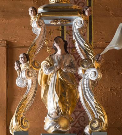 Zdjęcie nr 2: Feretron składa się z wolnostojącej, pełnoplastycznej rzeźby Matki Boskiej Niepokalanej stojącej na postumencie oraz fazowanym i profilowanym cokole, ponad którą wznosi się baldachim wsparty na czterech, spiętrzonych esowato wygiętych i srebrzonych zdwojonych wolutach. Woluty zdobią stylizowane, złocone palmety oraz rzeźbione, uskrzydlone główki aniołków, umieszczone na ich łączeniu, a także przy płaskim baldachimie, na którym spoczywa drewniana poduszka z koroną zamknietą. Całopostaciowa figura Marii ujęta została frontalnie w lekkim kontrapoście z modlitewnie złożonymi rękami uniesionymi na wysokości piersi. Niepokalana prawą stopę wspiera o srebrny  półksiężyc leżący na złotym globie, opleciony srebrnym wężem, który w paszczy trzyma jabłko. Jej głowa o owalnej twarzy, długiej szyi i jasnej karnacji oraz rumianych policzkach, drobnym nosie i wydatnych ustach jest nieznacznie odchylona w prawo. Długie, brązowe włosy opadają na jej plecy, odsłaniając uszy. Wokół głowy znajduje się druciany nimb podtrzymujący siedem złoconych gwiazd. Podniebie baldachimu ozdobiono płaskorzeźbionym, srebrzonym i złoconym wyobrażeniem gołębicy Ducha Świętego na tle promieni. Twarze uskrzydlonych aniołków na podporach baldachimu charakteryzują się jasną karnacją, wysokimi czołami, okrągłymi i pulchnymi policzkami oraz wydatnymi ustami. Ich głowy otaczają krótkie, brązowe włosy. Na szczycie baldachimu ustawiono rzeźbioną i srebrzoną poduszkę dekorowaną złocistym galonem i chwostami na rogach. Spoczywa na niej drewniana i złocona korona zamknięta.  