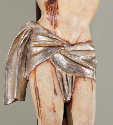 Zdjęcie nr 5: Na krucyfiksie przedstawiono ściętą z tyłu rzeźbioną figurę ukrzyżowanego Chrystusa. Jego ramiona ukazane zostały w lekkim zwisie, zaś układ ciała przyjmuje kształt litery „T”. Postać Ukrzyżowanego charakteryzuje się wydłużonymi proporcjami o wyraźnie zarysowanych mięśniach kończyn oraz zapadłą klatką piersiową. Głowa Jezusa jest przechylona na prawe ramię. Jego twarz jest pociągła o długim, prostym nosie, przymkniętych oczach i ustach wygiętych z kącikami skierowanymi do dołu. Okala ją brązowy zarost o szpicowatym zakończeniu. Włosy Zbawiciela są długie, kształtowane w falujące pukle, z pasmem opadającym na prawe ramię. Na głowie Jezus ma koronę cierniową, spod której spływają na twarz strugi krwi. Krew spływa także wzdłuż ramion, nóg i stóp. Na prawym boku zaznaczono broczącą ranę po włóczni. Ręce i nogi przytwierdzone są do krzyża za pomocą trzech gwoździ w kształcie czworobocznego graniastosłupa ściętego. Biodra Chrystusa przepasane zostały srebrzonym perizonium, przewiązanym na prawym biodrze ze zwisającym festonem wzdłuż prawego uda. Jest ono drapowane w szerokie fałdy wzdłuż przewiązania i na festonie. Ponad głową Zbawiciela widnieje późniejszy titulus z napisem „INRI”. Postać Chrystusa jest polichromowana, a perizonium srebrzone z widocznymi laserunkami.