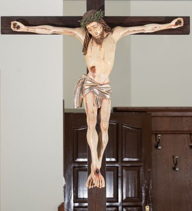 Zdjęcie nr 2: Na krucyfiksie przedstawiono ściętą z tyłu rzeźbioną figurę ukrzyżowanego Chrystusa. Jego ramiona ukazane zostały w lekkim zwisie, zaś układ ciała przyjmuje kształt litery „T”. Postać Ukrzyżowanego charakteryzuje się wydłużonymi proporcjami o wyraźnie zarysowanych mięśniach kończyn oraz zapadłą klatką piersiową. Głowa Jezusa jest przechylona na prawe ramię. Jego twarz jest pociągła o długim, prostym nosie, przymkniętych oczach i ustach wygiętych z kącikami skierowanymi do dołu. Okala ją brązowy zarost o szpicowatym zakończeniu. Włosy Zbawiciela są długie, kształtowane w falujące pukle, z pasmem opadającym na prawe ramię. Na głowie Jezus ma koronę cierniową, spod której spływają na twarz strugi krwi. Krew spływa także wzdłuż ramion, nóg i stóp. Na prawym boku zaznaczono broczącą ranę po włóczni. Ręce i nogi przytwierdzone są do krzyża za pomocą trzech gwoździ w kształcie czworobocznego graniastosłupa ściętego. Biodra Chrystusa przepasane zostały srebrzonym perizonium, przewiązanym na prawym biodrze ze zwisającym festonem wzdłuż prawego uda. Jest ono drapowane w szerokie fałdy wzdłuż przewiązania i na festonie. Ponad głową Zbawiciela widnieje późniejszy titulus z napisem „INRI”. Postać Chrystusa jest polichromowana, a perizonium srebrzone z widocznymi laserunkami.