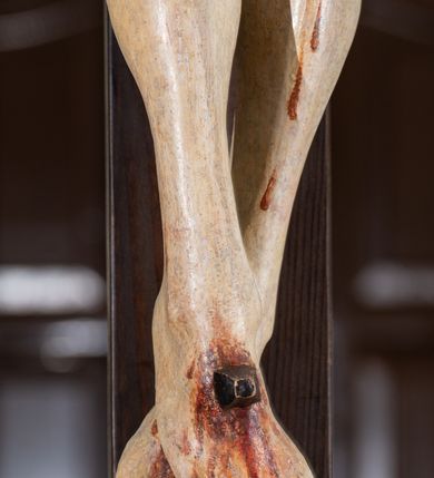 Zdjęcie nr 6: Na krucyfiksie przedstawiono ściętą z tyłu rzeźbioną figurę ukrzyżowanego Chrystusa. Jego ramiona ukazane zostały w lekkim zwisie, zaś układ ciała przyjmuje kształt litery „T”. Postać Ukrzyżowanego charakteryzuje się wydłużonymi proporcjami o wyraźnie zarysowanych mięśniach kończyn oraz zapadłą klatką piersiową. Głowa Jezusa jest przechylona na prawe ramię. Jego twarz jest pociągła o długim, prostym nosie, przymkniętych oczach i ustach wygiętych z kącikami skierowanymi do dołu. Okala ją brązowy zarost o szpicowatym zakończeniu. Włosy Zbawiciela są długie, kształtowane w falujące pukle, z pasmem opadającym na prawe ramię. Na głowie Jezus ma koronę cierniową, spod której spływają na twarz strugi krwi. Krew spływa także wzdłuż ramion, nóg i stóp. Na prawym boku zaznaczono broczącą ranę po włóczni. Ręce i nogi przytwierdzone są do krzyża za pomocą trzech gwoździ w kształcie czworobocznego graniastosłupa ściętego. Biodra Chrystusa przepasane zostały srebrzonym perizonium, przewiązanym na prawym biodrze ze zwisającym festonem wzdłuż prawego uda. Jest ono drapowane w szerokie fałdy wzdłuż przewiązania i na festonie. Ponad głową Zbawiciela widnieje późniejszy titulus z napisem „INRI”. Postać Chrystusa jest polichromowana, a perizonium srebrzone z widocznymi laserunkami.