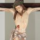 Zdjęcie nr 1: Na krucyfiksie przedstawiono ściętą z tyłu rzeźbioną figurę ukrzyżowanego Chrystusa. Jego ramiona ukazane zostały w lekkim zwisie, zaś układ ciała przyjmuje kształt litery „T”. Postać Ukrzyżowanego charakteryzuje się wydłużonymi proporcjami o wyraźnie zarysowanych mięśniach kończyn oraz zapadłą klatką piersiową. Głowa Jezusa jest przechylona na prawe ramię. Jego twarz jest pociągła o długim, prostym nosie, przymkniętych oczach i ustach wygiętych z kącikami skierowanymi do dołu. Okala ją brązowy zarost o szpicowatym zakończeniu. Włosy Zbawiciela są długie, kształtowane w falujące pukle, z pasmem opadającym na prawe ramię. Na głowie Jezus ma koronę cierniową, spod której spływają na twarz strugi krwi. Krew spływa także wzdłuż ramion, nóg i stóp. Na prawym boku zaznaczono broczącą ranę po włóczni. Ręce i nogi przytwierdzone są do krzyża za pomocą trzech gwoździ w kształcie czworobocznego graniastosłupa ściętego. Biodra Chrystusa przepasane zostały srebrzonym perizonium, przewiązanym na prawym biodrze ze zwisającym festonem wzdłuż prawego uda. Jest ono drapowane w szerokie fałdy wzdłuż przewiązania i na festonie. Ponad głową Zbawiciela widnieje późniejszy titulus z napisem „INRI”. Postać Chrystusa jest polichromowana, a perizonium srebrzone z widocznymi laserunkami.