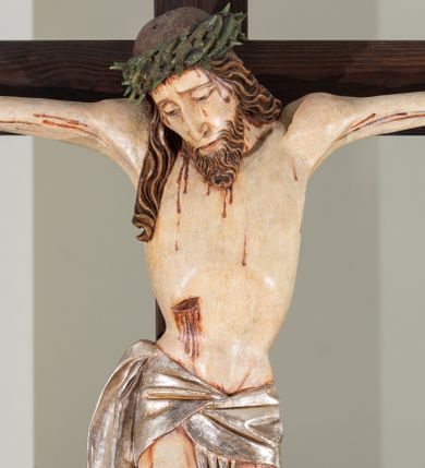 Zdjęcie nr 1: Na krucyfiksie przedstawiono ściętą z tyłu rzeźbioną figurę ukrzyżowanego Chrystusa. Jego ramiona ukazane zostały w lekkim zwisie, zaś układ ciała przyjmuje kształt litery „T”. Postać Ukrzyżowanego charakteryzuje się wydłużonymi proporcjami o wyraźnie zarysowanych mięśniach kończyn oraz zapadłą klatką piersiową. Głowa Jezusa jest przechylona na prawe ramię. Jego twarz jest pociągła o długim, prostym nosie, przymkniętych oczach i ustach wygiętych z kącikami skierowanymi do dołu. Okala ją brązowy zarost o szpicowatym zakończeniu. Włosy Zbawiciela są długie, kształtowane w falujące pukle, z pasmem opadającym na prawe ramię. Na głowie Jezus ma koronę cierniową, spod której spływają na twarz strugi krwi. Krew spływa także wzdłuż ramion, nóg i stóp. Na prawym boku zaznaczono broczącą ranę po włóczni. Ręce i nogi przytwierdzone są do krzyża za pomocą trzech gwoździ w kształcie czworobocznego graniastosłupa ściętego. Biodra Chrystusa przepasane zostały srebrzonym perizonium, przewiązanym na prawym biodrze ze zwisającym festonem wzdłuż prawego uda. Jest ono drapowane w szerokie fałdy wzdłuż przewiązania i na festonie. Ponad głową Zbawiciela widnieje późniejszy titulus z napisem „INRI”. Postać Chrystusa jest polichromowana, a perizonium srebrzone z widocznymi laserunkami.