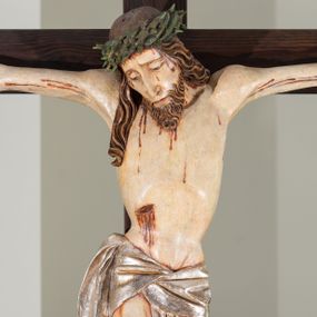 Zdjęcie nr 1: Na krucyfiksie przedstawiono ściętą z tyłu rzeźbioną figurę ukrzyżowanego Chrystusa. Jego ramiona ukazane zostały w lekkim zwisie, zaś układ ciała przyjmuje kształt litery „T”. Postać Ukrzyżowanego charakteryzuje się wydłużonymi proporcjami o wyraźnie zarysowanych mięśniach kończyn oraz zapadłą klatką piersiową. Głowa Jezusa jest przechylona na prawe ramię. Jego twarz jest pociągła o długim, prostym nosie, przymkniętych oczach i ustach wygiętych z kącikami skierowanymi do dołu. Okala ją brązowy zarost o szpicowatym zakończeniu. Włosy Zbawiciela są długie, kształtowane w falujące pukle, z pasmem opadającym na prawe ramię. Na głowie Jezus ma koronę cierniową, spod której spływają na twarz strugi krwi. Krew spływa także wzdłuż ramion, nóg i stóp. Na prawym boku zaznaczono broczącą ranę po włóczni. Ręce i nogi przytwierdzone są do krzyża za pomocą trzech gwoździ w kształcie czworobocznego graniastosłupa ściętego. Biodra Chrystusa przepasane zostały srebrzonym perizonium, przewiązanym na prawym biodrze ze zwisającym festonem wzdłuż prawego uda. Jest ono drapowane w szerokie fałdy wzdłuż przewiązania i na festonie. Ponad głową Zbawiciela widnieje późniejszy titulus z napisem „INRI”. Postać Chrystusa jest polichromowana, a perizonium srebrzone z widocznymi laserunkami.