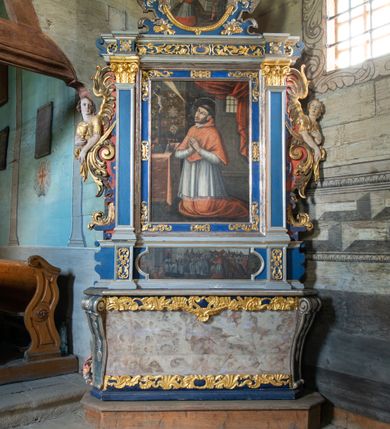 Zdjęcie nr 2: Retabulum św. Karola Boromeusza stanowi nastawę ze zwieńczeniem w formie architektonicznej, jednokondygnacyjną i jednoosiową. Wsparte jest na ośmiobocznej mensie spoczywającej na drewnianym stipesie w formie sarkofagowej, usytuowanym na niskim cokole. Boki stipesu ujmują złocone spływy wolutowe. Malowane antepedium ujęte jest przy górnej i dolnej krawędzi przez dwa pozłacane pasy z motywem małżowinowo-chrząstkowym. Predella retabulum w formie cokołu zawiera podłużne pole o nieregularnym kształcie z przedstawieniem Pokutnej procesji św. Karola Boromeusza w Mediolanie, ujęte po bokach dwoma prostokątnymi cokolikami, na których wspierają się pilastry retabulum. Retabulum jest oddzielone od predelli wąskim, spiętrzonym gzymsem, częściowo pozłacanym. Jego część środkową flankują dwa pilastry z głowicami kompozytowymi, wsparte na prostopadłościennych bazach, w których umieszczono prostokątne płyciny z motywem roślinnym. W centralnej części retabulum umieszczono obraz w formie stojącego prostokąta z przedstawieniem Modlitwy św. Karola Boromeusza. Po bokach nastawy znajdują się dwa uszaki uformowane z wici roślinno-kwiatowej, przedzielonej pośrodku płaskorzeźbionymi przedstawieniami aniołów. Ich zwrócone na zewnątrz sylwetki zostały wkomponowane motywy wolutowe. Anioł po lewej trzyma w prawej dłoni kielich mszalny, a anioł po prawej stronie ołtarza ma wzrok zwrócony w górę. Lewą, zgiętą w łokciu rękę trzyma na piersi, prawą ma wyprostowaną. Obraz jest ujęty w ramę o profilowanych bokach, zdobionych luźnymi motywami roślinnymi w formie stylizowanych liści akantu. Ponad nią widoczne jest profilowane belkowanie, załamane na osi pilastrów, wzbogacone na środku fryzem z motywami roślinnymi. W zwieńczeniu ołtarza umieszczono owalny obraz ze sceną Zamachu na św. Karola Boromeusza, ujęty ornamentem kartuszowym z elementami akantowymi i wolutowymi.