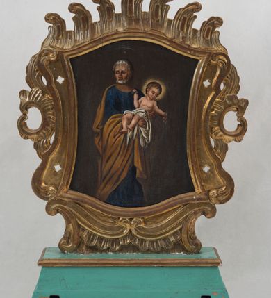 Zdjęcie nr 2: Pole obrazowe feretronu uzyskało kształt nieregularny. Ujmuje je rzeźbiona, wklęsło-wypukła profilowana rama, otoczona spływami wolutowymi oraz motywami rocaille. Niski, fazowany postument, do którego przymocowano górną część feretronu, przepruto dwoma kwadratowymi otworami umożliwiającymi włożenie drążków do noszenia. Na awersie feretronu w polu obrazowym przedstawiono Matkę Boską z Dzieciątkiem. Maria została ukazana frontalnie, w półpostaci. Na lewym ramieniu trzyma Dzieciątko, któremu prawą ręką podaje jabłko. Madonna ma jasną karnację z rumianymi policzkami. Jej twarz charakteryzuje wydatny, prosty nos, duże, ciemne oczy oraz wyraziste usta. Na głowie ma ciemnogranatowy maforion podbity zielenią, lamowany złotą taśmą dekorowaną naśladownictwem kameryzacji. Spod maforionu wokół głowy widać białą chustę. Maforion jest spięty pod szyją złotą fibulą w kształcie stylizowanego kwiatu. Okrywa on ramiona Marii odsłaniając jednocześnie na piersi i pod prawą ręką fragmenty fioletowej sukni spodniej, także lamowanej złotą taśmą z imitacją kameryzacji. Na prawym ramieniu maforion ozdobiony jest złotą, ośmioramienną gwiazdą w otoku. Matka Boska ma na lekko pochylonej w lewo głowie złotą, zamkniętą koronę, zwieńczoną globem z krzyżykiem. Obręcz i pałąki korony zdobione są malowanymi kamieniami szlachetnymi. Siedzący na lewym ramieniu mały Jezus ujęty został w trzech czwartych. W lewej ręce trzyma kodeks Ewangelii, a prawą sięga po jabłko trzymane przez Matkę w prawej ręce. Dzieciątko o rumianych policzkach ma jasną karnację. Jego twarz jest owalna. Charakteryzują ją duże, ciemne oczy, wydatny nos i czerwone usta. Na głowie ma krótkie, brązowe włosy z charakterystycznymi lokami i kosmykiem wychylającym się spod korony na czole. Mały Jezus ubrany jest w lamowaną złotą taśmą z imitacją kameryzacji szkarłatną tunikę, przewiązaną taką samą taśmą w pasie. Spod niej widoczne są częściowo obie jego stopy obute w sandały. Jezus ma na głowie zamkniętą złotą koronę, zwieńczoną krzyżykiem. Zarówno obręcz, jak i pałąki dekorowane są malarską imitacją kameryzacji. Postacie zostały przedstawione na brązowym tle, rozjaśnionym świetlistymi nimbami widocznymi wokół głowy Madonny i Dzieciątka. Na rewersie feretronu przedstawiono św. Józefa z Dzieciątkiem na rękach. Józef został ukazany stojąc w dwóch trzecich postaci. Ujęty został w trzech czwartych w prawo. Jest to starszy mężczyzna z wyraźnie zwróconą głową w prawą stronę. Nad jego głową widać nieznacznie zaznaczony nimb talerzowy z gwiazdą w środku. Ma siwe włosy i taki sam krótki zarost. Jego twarz jest pociągła o śniadej karnacji z wydatnym nosem, ustami i szeroko otwartymi niebieskimi oczami. Ma na sobie ciemnoniebieską tunikę oraz osuwający się z ramion ciemnoochrowy płaszcz zakrywający zgięte w łokciach ręce, którymi podtrzymuje Dzieciątko. Mały Jezus został ujęty nago w trzech czwartych w lewo z wyraźnie zwróconą głową w tę samą stronę. Odchyla się od Józefa, trzymając w uniesionych rączkach drobne gałązki różanego krzewu. Ma jasną karnację. Jego twarz charakteryzuje wysokie czoło, częściowo zasłonięte przez krótkie, pofalowane włosy. Dzieciątko ma kształtnie malowany nos i usta oraz deltę nosową z lekko przymkniętymi oczami. Biodra Pana Jezusa okrywa biała chusta, odsłaniająca jego nóżki, spod której widać dłonie Józefa. Głowę małego Jezusa otacza świetlisty nimb.