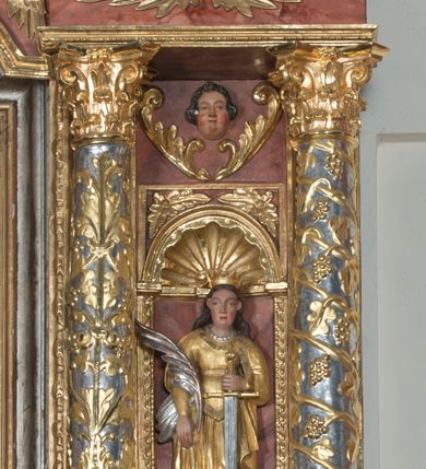 Zdjęcie nr 12: Ołtarz główny w Głębowicach otrzymał strukturę przyściennego, trójosiowego retabulum architektonicznego o dwóch kondygnacjach. Zaopatrzono go w predellę i zwieńczenie oraz dwa uszaki z boku zdobione herbem Starykoń i Jelita. Wisząc na ścianie wspiera się na dwóch masywnych, krótkich półkolumnach flankujących wejście do zakrystii. W środkowej części predelli widoczne jest wycięcie. Predellę po bokach ujmuje para konsoli wspierających wyłamujący się ponad nimi gzyms. Osie części środkowej nastawy ujęte są parami wolnostojących kolumn o kapitelach korynckich wspierających wyłamujące się ponad nimi belkowanie. Pole części środkowej nastawy ma kształt stojącego prostokąta zamkniętego nadwieszonym łukiem odcinkowym. Z kolei pola osi bocznych są artykułowane parą zamkniętych półkoliście niszy. Druga kondygnacja retabulum, będąca zarazem jego zwieńczeniem, została ujęta parą herm wspierających belkowanie przybierające formę łuku trójbocznego z ośmioboku. Po bokach widoczna jest para półkolistych, przerwanych przyczółków. Boki predelli ujęte zostały ulistnionymi wolutami, dodatkowo zdobionymi stylizowanymi motywami roślinno-kwiatowymi oraz motywem zwisu tkaniny z supłem. Konsole zdobione są liśćmi akantu oraz parą uskrzydlonych główek anielskich ze zwisami w formie pojedynczych kampanull. Po bokach predelli, na przedłużeniu zewnętrznych kolumn nastawy, podwieszono parę szyszek. Trzony kolumn zostały ozdobione w dolnej partii płaskorzeźbionymi, uskrzydlonymi główkami anielskimi, w górnej zewnętrzne – wicią winnej latorośli, zaś wewnętrzne motywem kandelabru. Osi boczne nastawy zostały ozdobione w dolnej partii ornamnetyką roślinną i parą główek anielskich ujętych motywami roślinnymi. Nisze dolnej kondygnacji wypełniono konchami, zdobionymi motywem ulistnionych rozetek. Z kolei trójboczne z ośmioboku konsole zostały ozdobione stylizowanymi liśćmi. W partii fryzu widoczna jest para mniejszych i większych uskrzydlonych główek anielskich. Drugą kondygnację - zwieńczenie, zostało ujęte parą herm z sylwetkami kobiecymi oraz parą uszaków o fantazyjnym wykroju z motywem maski lunarnej. Także fantazyjną formę z motywem głowy gryfa trzymającego w dziobie szarfę uzyskały uszaki w dolnej kondygnacji. W ich centrum widoczne są dwa owalne medaliony z herbem Starykoń i Jelita. W części środkowej nastawy znajduje się obraz Franciszka Lekszyckiego "Matka Boska Szkaplerzna wręcza szkaplerz św. Szymonowi Stockowi". Na zasuwie obraz "Matka Boska Różańcowa". Po bokach obrazu w niszach flankowanych kolumnami ustawiono rzeźbione wyobrażenie św. Marii Magdaleny i św. Katarzyny Aleksandryjskiej. W centrum drugiej kondygnacji-zwieńczeniu znajduje się obraz "Św. Maria Magdalena przed Ukrzyżowanym Chrystusem". Po bokach, na przyczółkach znajduje się para rzeźbionych stojących aniołów trzymających w dłoniach kartusze o fantazyjnych wykrojach. W kartuszu z prawej strony widnieje napis: "konserwacja 1973, 2016", a z lewej: "sancta scapularis". Na szczycie ołtarza trzy rzeźbione przedstawienia - bo bokach św. Piotra i św. Jana Ewangelisty, a po środku św. Krzysztofa. Struktura nastawy jest marmoryzowana w odcieniach różu i ceglastej czerwieni. Partie detalu architektonicznego i ornamentyka są złocone i srebrzone.