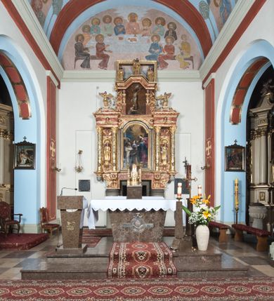 Zdjęcie nr 2: Ołtarz główny w Głębowicach otrzymał strukturę przyściennego, trójosiowego retabulum architektonicznego o dwóch kondygnacjach. Zaopatrzono go w predellę i zwieńczenie oraz dwa uszaki z boku zdobione herbem Starykoń i Jelita. Wisząc na ścianie wspiera się na dwóch masywnych, krótkich półkolumnach flankujących wejście do zakrystii. W środkowej części predelli widoczne jest wycięcie. Predellę po bokach ujmuje para konsoli wspierających wyłamujący się ponad nimi gzyms. Osie części środkowej nastawy ujęte są parami wolnostojących kolumn o kapitelach korynckich wspierających wyłamujące się ponad nimi belkowanie. Pole części środkowej nastawy ma kształt stojącego prostokąta zamkniętego nadwieszonym łukiem odcinkowym. Z kolei pola osi bocznych są artykułowane parą zamkniętych półkoliście niszy. Druga kondygnacja retabulum, będąca zarazem jego zwieńczeniem, została ujęta parą herm wspierających belkowanie przybierające formę łuku trójbocznego z ośmioboku. Po bokach widoczna jest para półkolistych, przerwanych przyczółków. Boki predelli ujęte zostały ulistnionymi wolutami, dodatkowo zdobionymi stylizowanymi motywami roślinno-kwiatowymi oraz motywem zwisu tkaniny z supłem. Konsole zdobione są liśćmi akantu oraz parą uskrzydlonych główek anielskich ze zwisami w formie pojedynczych kampanull. Po bokach predelli, na przedłużeniu zewnętrznych kolumn nastawy, podwieszono parę szyszek. Trzony kolumn zostały ozdobione w dolnej partii płaskorzeźbionymi, uskrzydlonymi główkami anielskimi, w górnej zewnętrzne – wicią winnej latorośli, zaś wewnętrzne motywem kandelabru. Osi boczne nastawy zostały ozdobione w dolnej partii ornamnetyką roślinną i parą główek anielskich ujętych motywami roślinnymi. Nisze dolnej kondygnacji wypełniono konchami, zdobionymi motywem ulistnionych rozetek. Z kolei trójboczne z ośmioboku konsole zostały ozdobione stylizowanymi liśćmi. W partii fryzu widoczna jest para mniejszych i większych uskrzydlonych główek anielskich. Drugą kondygnację - zwieńczenie, zostało ujęte parą herm z sylwetkami kobiecymi oraz parą uszaków o fantazyjnym wykroju z motywem maski lunarnej. Także fantazyjną formę z motywem głowy gryfa trzymającego w dziobie szarfę uzyskały uszaki w dolnej kondygnacji. W ich centrum widoczne są dwa owalne medaliony z herbem Starykoń i Jelita. W części środkowej nastawy znajduje się obraz Franciszka Lekszyckiego "Matka Boska Szkaplerzna wręcza szkaplerz św. Szymonowi Stockowi". Na zasuwie obraz "Matka Boska Różańcowa". Po bokach obrazu w niszach flankowanych kolumnami ustawiono rzeźbione wyobrażenie św. Marii Magdaleny i św. Katarzyny Aleksandryjskiej. W centrum drugiej kondygnacji-zwieńczeniu znajduje się obraz "Św. Maria Magdalena przed Ukrzyżowanym Chrystusem". Po bokach, na przyczółkach znajduje się para rzeźbionych stojących aniołów trzymających w dłoniach kartusze o fantazyjnych wykrojach. W kartuszu z prawej strony widnieje napis: "konserwacja 1973, 2016", a z lewej: "sancta scapularis". Na szczycie ołtarza trzy rzeźbione przedstawienia - bo bokach św. Piotra i św. Jana Ewangelisty, a po środku św. Krzysztofa. Struktura nastawy jest marmoryzowana w odcieniach różu i ceglastej czerwieni. Partie detalu architektonicznego i ornamentyka są złocone i srebrzone.