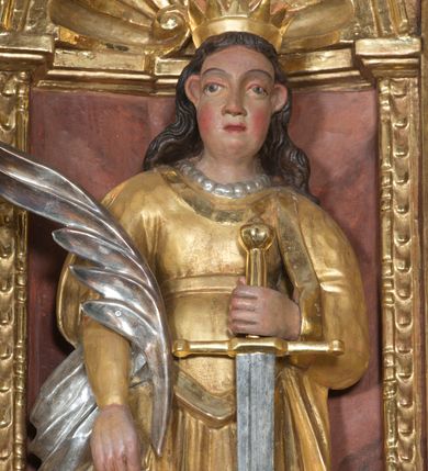 Zdjęcie nr 3: Święta została przedstawiona frontalnie w całej postaci w nieznacznym kontrapoście. Ma owalną twarz o sumarycznym modelunku rysów i rumianych policzkach. Jej długie, lekko falowane, ciemnobrązowe włosy, założone za wydatne uszy, opadają na plecy. Jest ubrana w długą, złoconą suknię z gorsetem oraz osłaniający ramiona, złocony płaszcz podwinięty pod prawą ręką. Na głowie ma otwartą, złoconą koronę, a na szyi sznur pereł. W prawej ręce opuszczonej wzdłuż ciała trzyma srebrzoną gałązkę palmową. Uniesioną na wysokość talii lewą ręką wspiera się o miecz o złoconej rękojeści i srebrzonym ostrzu. Układ draperii dolnej części szat z lewej strony jest nieznacznie odsunięty, co sugeruje, że w tym miejscu najprawdopodobniej znajdował się fragment zębatego koła. 