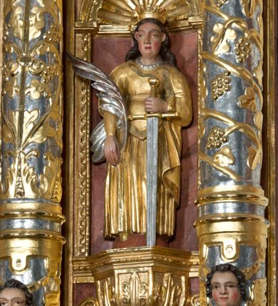 Zdjęcie nr 1: Święta została przedstawiona frontalnie w całej postaci w nieznacznym kontrapoście. Ma owalną twarz o sumarycznym modelunku rysów i rumianych policzkach. Jej długie, lekko falowane, ciemnobrązowe włosy, założone za wydatne uszy, opadają na plecy. Jest ubrana w długą, złoconą suknię z gorsetem oraz osłaniający ramiona, złocony płaszcz podwinięty pod prawą ręką. Na głowie ma otwartą, złoconą koronę, a na szyi sznur pereł. W prawej ręce opuszczonej wzdłuż ciała trzyma srebrzoną gałązkę palmową. Uniesioną na wysokość talii lewą ręką wspiera się o miecz o złoconej rękojeści i srebrzonym ostrzu. Układ draperii dolnej części szat z lewej strony jest nieznacznie odsunięty, co sugeruje, że w tym miejscu najprawdopodobniej znajdował się fragment zębatego koła. 