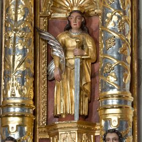 Zdjęcie nr 1: Święta została przedstawiona frontalnie w całej postaci w nieznacznym kontrapoście. Ma owalną twarz o sumarycznym modelunku rysów i rumianych policzkach. Jej długie, lekko falowane, ciemnobrązowe włosy, założone za wydatne uszy, opadają na plecy. Jest ubrana w długą, złoconą suknię z gorsetem oraz osłaniający ramiona, złocony płaszcz podwinięty pod prawą ręką. Na głowie ma otwartą, złoconą koronę, a na szyi sznur pereł. W prawej ręce opuszczonej wzdłuż ciała trzyma srebrzoną gałązkę palmową. Uniesioną na wysokość talii lewą ręką wspiera się o miecz o złoconej rękojeści i srebrzonym ostrzu. Układ draperii dolnej części szat z lewej strony jest nieznacznie odsunięty, co sugeruje, że w tym miejscu najprawdopodobniej znajdował się fragment zębatego koła. 