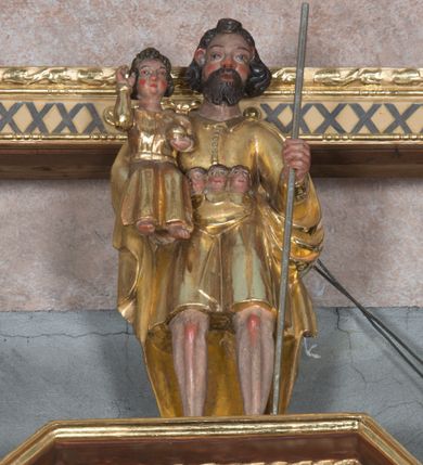 Zdjęcie nr 1: Rzeźba ukazuje całopostaciową figurę stojącego frontalnie na nieznacznie rozstawionych nogach św. Krzysztofa niosącego małego Chrystusa oraz innych podróżnych. Twarz świętego charakteryzuje sumaryczny modelunek rysów oraz krótki, dwudzielny zarost. Jego falujące, brązowe włosy sięgające karku są założone za wydatne uszy. Nad czołem widoczny jest charakterystyczny pukiel włosów. Święty jest ubrany w sięgającą kolan, złoconą tunikę, zapinaną od góry na guziki i osłaniający plecy, podwinięty pod lewą rękę złocony płaszcz. Tunika przepasana jest w pasie symboliczną torbą, z której wychylają się trzy niewielkie ludzkie głowy. Święty Krzysztof lewą ręką wspiera się na długiej, srebrzonej lasce. Na jego prawym przedramieniu siedzi frontalnie Dzieciątko Jezus. Mały Chrystus ubrany jest w długą, złoconą tunikę, spod której widać jego stopy. Ma pociągłą twarz o wydatnych rumianych policzkach, okoloną puklowanymi włosami sięgającymi szyi. W lewej ręce trzyma złocony glob, prawą zaś unosi w geście błogosławieństwa.