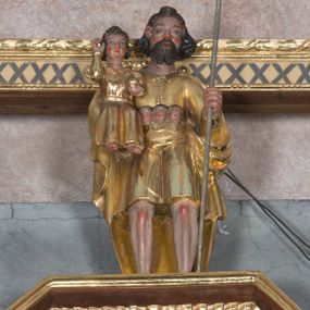 Zdjęcie nr 1: Rzeźba ukazuje całopostaciową figurę stojącego frontalnie na nieznacznie rozstawionych nogach św. Krzysztofa niosącego małego Chrystusa oraz innych podróżnych. Twarz świętego charakteryzuje sumaryczny modelunek rysów oraz krótki, dwudzielny zarost. Jego falujące, brązowe włosy sięgające karku są założone za wydatne uszy. Nad czołem widoczny jest charakterystyczny pukiel włosów. Święty jest ubrany w sięgającą kolan, złoconą tunikę, zapinaną od góry na guziki i osłaniający plecy, podwinięty pod lewą rękę złocony płaszcz. Tunika przepasana jest w pasie symboliczną torbą, z której wychylają się trzy niewielkie ludzkie głowy. Święty Krzysztof lewą ręką wspiera się na długiej, srebrzonej lasce. Na jego prawym przedramieniu siedzi frontalnie Dzieciątko Jezus. Mały Chrystus ubrany jest w długą, złoconą tunikę, spod której widać jego stopy. Ma pociągłą twarz o wydatnych rumianych policzkach, okoloną puklowanymi włosami sięgającymi szyi. W lewej ręce trzyma złocony glob, prawą zaś unosi w geście błogosławieństwa.