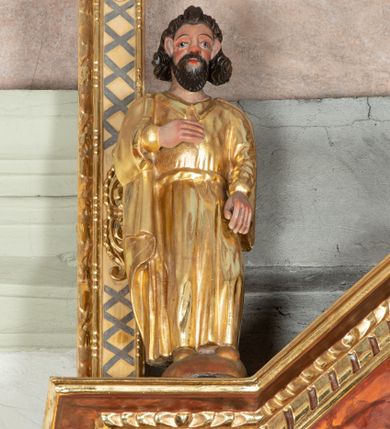 Zdjęcie nr 1: Rzeźbiona figura ukazuje całopostaciowe wyobrażenie stojącego frontalnie św. Jana Apostoła. Święty ma owalną, szeroką twarz o sumarycznym modelunku rysów i rumianych policzkach z krótkim, ciemnobrązowym zarostem. Jego brązowe włosy układają się w pukle założone za wydatne uszy. Apostoł ubrany jest w długą, złoconą przewiązaną w pasie tunikę z kołnierzykiem oraz złocony płaszcz. Prawą rękę wspiera na piersiach. Pierwotnie trzymał w niej kielich.