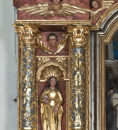 Zdjęcie nr 3: Święta Maria Magdalena, identyfikowana przez trzymaną ludzką czaszkę, została ukazana w całej postaci w nieznacznym kontrapoście. Rzeźba ma lekko zwróconą w lewo głowę. Święta ma owalną twarz o sumarycznym modelunku rysów i wyraźnie rumianych policzkach. Jej długie, lekko falowane, ciemnobrązowe włosy opadające na ramiona są założone za wydatne uszy. Maria Magdalena ubrana jest w długą, złoconą suknię z gorsetem oraz złocony, spięty pod szyją płaszcz. Na szyi widoczny jest krótki sznur pereł. Pod prawą pachą trzyma w uniesionej na wysokości talii prawej ręce ludzką czaszkę. Lewą zaś rękę wspiera na udzie. Figura nieznacznie ścięta z tyłu umieszczona jest w niszy pomiędzy dwiema kolumnami z prawej strony retabulum.