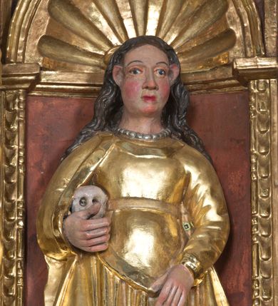 Zdjęcie nr 2: Święta Maria Magdalena, identyfikowana przez trzymaną ludzką czaszkę, została ukazana w całej postaci w nieznacznym kontrapoście. Rzeźba ma lekko zwróconą w lewo głowę. Święta ma owalną twarz o sumarycznym modelunku rysów i wyraźnie rumianych policzkach. Jej długie, lekko falowane, ciemnobrązowe włosy opadające na ramiona są założone za wydatne uszy. Maria Magdalena ubrana jest w długą, złoconą suknię z gorsetem oraz złocony, spięty pod szyją płaszcz. Na szyi widoczny jest krótki sznur pereł. Pod prawą pachą trzyma w uniesionej na wysokości talii prawej ręce ludzką czaszkę. Lewą zaś rękę wspiera na udzie. Figura nieznacznie ścięta z tyłu umieszczona jest w niszy pomiędzy dwiema kolumnami z prawej strony retabulum.
