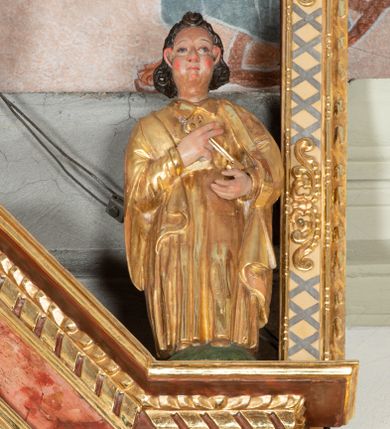 Zdjęcie nr 1: Święty Piotr został przedstawiony w całej postaci frontalnie. Ma owalną, szeroką twarz o sumarycznym modelunku rysów i wyraźnie rumianych policzkach. Jego ciemnobrązowe włosy sięgające szyi są założone za wydatne uszy. Święty ubrany jest w długą, złoconą przewiązaną w pasie tunikę oraz złocony płaszcz. W uniesionych na wysokości klatki piersiowej rękach trzyma dwa klucze, złoty i srebrny. 