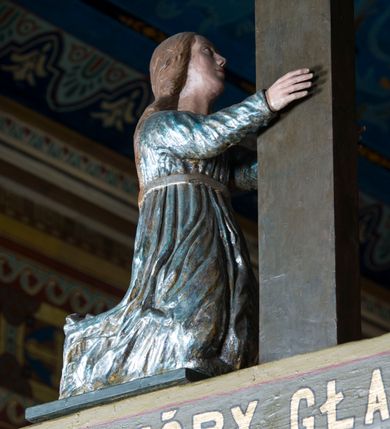Zdjęcie nr 9: Grupa Ukrzyżowania składa się z pełnoplastycznych, całopostaciowych figur ustawionych na belce tęczowej. Pośrodku zamocowano polichromowany na ciemnobrązowo krzyż z realistycznie rzeźbioną figurą Chrystusa o plastycznie opracowanym modelunku muskulatury ciała. Postać do krzyża przytwierdzono za pomocą trzech gwoździ. Zbawiciel ma głowę pochyloną i złożoną na prawym barku. Posiada szeroką, owalną twarz o sumarycznie opracowanych rysach, półprzymknięte oczy, lekko rozchylone usta i krótki, ciemnobrązowy zarost. Długie, ciemnobrązowe, falowane włosy opadają mu na plecy i na prawy bark. Na głowie znajduje się zielona, cierniowa korona, spod której ciekną stróżki krwi. Nieproporcjonalnie długie ramiona ułożone zostały w płytkim, U-kształtnym zwisie z zaznaczonymi ścięgnami i żyłami na przedramionach. Dłonie o zaciśniętych palcach przybito dwoma gwoździami, spod których ciekną strużki krwi. Klatkę piersiową ukształtowano z wyraziście zaznaczoną linią żeber i raną w boku ze strugami krwi. Na biodrach umiejscowiono krótkie, złocone perizonium z festonem przy prawym boku. Jego zwichrowane draperie zostały ułożone horyzontalnie. Chrystus ma zgięte w kolanach nogi z założoną prawą stopą na lewą przybite jednym gwoździem. W górnej partii pionowej belki krzyża zawieszono biały titulus w formie banderoli z napisem: "INRI". 
U stóp krzyża, po lewej stronie, na niskiej, prostopadłościennej podstawie, ustawiono rzeźbę klęczącej św. Marii Magdaleny. Święta jest zwrócona profilem w prawo. Jej owalną twarz charakteryzują sumarycznie opracowane rysy. Ma długie brązowe, lekko falowane włosy opadające na plecy. Ubrana jest w długą, srebrzystobłękitną, drobno fałdowaną suknię związaną srebrnym paskiem. Oburącz ujmuje pionową belkę krzyża. Tuż obok niej, na lewo, na niskiej, prostopadłościennej podstawie ustawiono figurę stojącej frontalnie Matki Boskiej Bolesnej. Jej nieznacznie odchyloną głowę charakteryzuje owalna, szeroka twarz o sumarycznie opracowanych rysach i wzroku skierowanym ku górze. Maria ubrana jest w długą srebrzyścieczerwoną suknię przewiązaną złotym paskiem i osłaniający głowę, plecy i prawe ramię, drobnofałdowany srebrzysty płaszcz. Ręce trzyma złożone w geście modlitwy. Po prawej stronie krzyża na niskiej, prostopadłościennej podstawie zamocowano stojącą figurę św. Jana Ewangelisty, ujętego frontalnie w nieznacznym kontrapoście. Święty ma lekko odchyloną do tyłu, zwróconą w trzech czwartych w lewo głowę. Charakteryzuje ją owalna, szeroka twarz o sumarycznie opracowanych rysach. Na plecy opadają mu długie, falowane, brązowe włosy. Jan ubrany jest w długą, srebrzystozieloną tunikę i przerzucony przez prawe ramię i podtrzymywany lewym przedramieniem złocistoochrowy płaszcz. Ręce trzyma skrzyżowane na piersiach. Z tyłu titulusa widnieje napis: Malował Jan i Ludwik, bracia Stankiewicze 1913. Napis widoczny jest także na górnej płaszczyźnie z tyłu krzyża: Malował 1913. 
Belka tęczowa jest prostopadłościenna, o profilowanych dolnych narożach. Polichromowano ją w odcieniach zieleni, czerwieni i żółci. Na środku widnieje flankowany parą złoconych krzyżyków złocony z przodu napis: + OTO BARANEK BOŻY KTÓRY GŁADZI GRZECHY ŚWIATA +; z tyłu: RENOVATUM A[nno] D[omini] 1913 + 1953 + 1983 + 2013