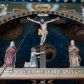 Zdjęcie nr 1: Grupa Ukrzyżowania składa się z pełnoplastycznych, całopostaciowych figur ustawionych na belce tęczowej. Pośrodku zamocowano polichromowany na ciemnobrązowo krzyż z realistycznie rzeźbioną figurą Chrystusa o plastycznie opracowanym modelunku muskulatury ciała. Postać do krzyża przytwierdzono za pomocą trzech gwoździ. Zbawiciel ma głowę pochyloną i złożoną na prawym barku. Posiada szeroką, owalną twarz o sumarycznie opracowanych rysach, półprzymknięte oczy, lekko rozchylone usta i krótki, ciemnobrązowy zarost. Długie, ciemnobrązowe, falowane włosy opadają mu na plecy i na prawy bark. Na głowie znajduje się zielona, cierniowa korona, spod której ciekną stróżki krwi. Nieproporcjonalnie długie ramiona ułożone zostały w płytkim, U-kształtnym zwisie z zaznaczonymi ścięgnami i żyłami na przedramionach. Dłonie o zaciśniętych palcach przybito dwoma gwoździami, spod których ciekną strużki krwi. Klatkę piersiową ukształtowano z wyraziście zaznaczoną linią żeber i raną w boku ze strugami krwi. Na biodrach umiejscowiono krótkie, złocone perizonium z festonem przy prawym boku. Jego zwichrowane draperie zostały ułożone horyzontalnie. Chrystus ma zgięte w kolanach nogi z założoną prawą stopą na lewą przybite jednym gwoździem. W górnej partii pionowej belki krzyża zawieszono biały titulus w formie banderoli z napisem: "INRI". 
U stóp krzyża, po lewej stronie, na niskiej, prostopadłościennej podstawie, ustawiono rzeźbę klęczącej św. Marii Magdaleny. Święta jest zwrócona profilem w prawo. Jej owalną twarz charakteryzują sumarycznie opracowane rysy. Ma długie brązowe, lekko falowane włosy opadające na plecy. Ubrana jest w długą, srebrzystobłękitną, drobno fałdowaną suknię związaną srebrnym paskiem. Oburącz ujmuje pionową belkę krzyża. Tuż obok niej, na lewo, na niskiej, prostopadłościennej podstawie ustawiono figurę stojącej frontalnie Matki Boskiej Bolesnej. Jej nieznacznie odchyloną głowę charakteryzuje owalna, szeroka twarz o sumarycznie opracowanych rysach i wzroku skierowanym ku górze. Maria ubrana jest w długą srebrzyścieczerwoną suknię przewiązaną złotym paskiem i osłaniający głowę, plecy i prawe ramię, drobnofałdowany srebrzysty płaszcz. Ręce trzyma złożone w geście modlitwy. Po prawej stronie krzyża na niskiej, prostopadłościennej podstawie zamocowano stojącą figurę św. Jana Ewangelisty, ujętego frontalnie w nieznacznym kontrapoście. Święty ma lekko odchyloną do tyłu, zwróconą w trzech czwartych w lewo głowę. Charakteryzuje ją owalna, szeroka twarz o sumarycznie opracowanych rysach. Na plecy opadają mu długie, falowane, brązowe włosy. Jan ubrany jest w długą, srebrzystozieloną tunikę i przerzucony przez prawe ramię i podtrzymywany lewym przedramieniem złocistoochrowy płaszcz. Ręce trzyma skrzyżowane na piersiach. Z tyłu titulusa widnieje napis: Malował Jan i Ludwik, bracia Stankiewicze 1913. Napis widoczny jest także na górnej płaszczyźnie z tyłu krzyża: Malował 1913. 
Belka tęczowa jest prostopadłościenna, o profilowanych dolnych narożach. Polichromowano ją w odcieniach zieleni, czerwieni i żółci. Na środku widnieje flankowany parą złoconych krzyżyków złocony z przodu napis: + OTO BARANEK BOŻY KTÓRY GŁADZI GRZECHY ŚWIATA +; z tyłu: RENOVATUM A[nno] D[omini] 1913 + 1953 + 1983 + 2013