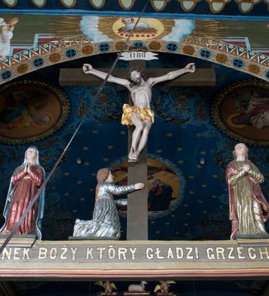 Zdjęcie nr 1: Grupa Ukrzyżowania składa się z pełnoplastycznych, całopostaciowych figur ustawionych na belce tęczowej. Pośrodku zamocowano polichromowany na ciemnobrązowo krzyż z realistycznie rzeźbioną figurą Chrystusa o plastycznie opracowanym modelunku muskulatury ciała. Postać do krzyża przytwierdzono za pomocą trzech gwoździ. Zbawiciel ma głowę pochyloną i złożoną na prawym barku. Posiada szeroką, owalną twarz o sumarycznie opracowanych rysach, półprzymknięte oczy, lekko rozchylone usta i krótki, ciemnobrązowy zarost. Długie, ciemnobrązowe, falowane włosy opadają mu na plecy i na prawy bark. Na głowie znajduje się zielona, cierniowa korona, spod której ciekną stróżki krwi. Nieproporcjonalnie długie ramiona ułożone zostały w płytkim, U-kształtnym zwisie z zaznaczonymi ścięgnami i żyłami na przedramionach. Dłonie o zaciśniętych palcach przybito dwoma gwoździami, spod których ciekną strużki krwi. Klatkę piersiową ukształtowano z wyraziście zaznaczoną linią żeber i raną w boku ze strugami krwi. Na biodrach umiejscowiono krótkie, złocone perizonium z festonem przy prawym boku. Jego zwichrowane draperie zostały ułożone horyzontalnie. Chrystus ma zgięte w kolanach nogi z założoną prawą stopą na lewą przybite jednym gwoździem. W górnej partii pionowej belki krzyża zawieszono biały titulus w formie banderoli z napisem: "INRI". 
U stóp krzyża, po lewej stronie, na niskiej, prostopadłościennej podstawie, ustawiono rzeźbę klęczącej św. Marii Magdaleny. Święta jest zwrócona profilem w prawo. Jej owalną twarz charakteryzują sumarycznie opracowane rysy. Ma długie brązowe, lekko falowane włosy opadające na plecy. Ubrana jest w długą, srebrzystobłękitną, drobno fałdowaną suknię związaną srebrnym paskiem. Oburącz ujmuje pionową belkę krzyża. Tuż obok niej, na lewo, na niskiej, prostopadłościennej podstawie ustawiono figurę stojącej frontalnie Matki Boskiej Bolesnej. Jej nieznacznie odchyloną głowę charakteryzuje owalna, szeroka twarz o sumarycznie opracowanych rysach i wzroku skierowanym ku górze. Maria ubrana jest w długą srebrzyścieczerwoną suknię przewiązaną złotym paskiem i osłaniający głowę, plecy i prawe ramię, drobnofałdowany srebrzysty płaszcz. Ręce trzyma złożone w geście modlitwy. Po prawej stronie krzyża na niskiej, prostopadłościennej podstawie zamocowano stojącą figurę św. Jana Ewangelisty, ujętego frontalnie w nieznacznym kontrapoście. Święty ma lekko odchyloną do tyłu, zwróconą w trzech czwartych w lewo głowę. Charakteryzuje ją owalna, szeroka twarz o sumarycznie opracowanych rysach. Na plecy opadają mu długie, falowane, brązowe włosy. Jan ubrany jest w długą, srebrzystozieloną tunikę i przerzucony przez prawe ramię i podtrzymywany lewym przedramieniem złocistoochrowy płaszcz. Ręce trzyma skrzyżowane na piersiach. Z tyłu titulusa widnieje napis: Malował Jan i Ludwik, bracia Stankiewicze 1913. Napis widoczny jest także na górnej płaszczyźnie z tyłu krzyża: Malował 1913. 
Belka tęczowa jest prostopadłościenna, o profilowanych dolnych narożach. Polichromowano ją w odcieniach zieleni, czerwieni i żółci. Na środku widnieje flankowany parą złoconych krzyżyków złocony z przodu napis: + OTO BARANEK BOŻY KTÓRY GŁADZI GRZECHY ŚWIATA +; z tyłu: RENOVATUM A[nno] D[omini] 1913 + 1953 + 1983 + 2013