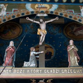 Zdjęcie nr 1: Grupa Ukrzyżowania składa się z pełnoplastycznych, całopostaciowych figur ustawionych na belce tęczowej. Pośrodku zamocowano polichromowany na ciemnobrązowo krzyż z realistycznie rzeźbioną figurą Chrystusa o plastycznie opracowanym modelunku muskulatury ciała. Postać do krzyża przytwierdzono za pomocą trzech gwoździ. Zbawiciel ma głowę pochyloną i złożoną na prawym barku. Posiada szeroką, owalną twarz o sumarycznie opracowanych rysach, półprzymknięte oczy, lekko rozchylone usta i krótki, ciemnobrązowy zarost. Długie, ciemnobrązowe, falowane włosy opadają mu na plecy i na prawy bark. Na głowie znajduje się zielona, cierniowa korona, spod której ciekną stróżki krwi. Nieproporcjonalnie długie ramiona ułożone zostały w płytkim, U-kształtnym zwisie z zaznaczonymi ścięgnami i żyłami na przedramionach. Dłonie o zaciśniętych palcach przybito dwoma gwoździami, spod których ciekną strużki krwi. Klatkę piersiową ukształtowano z wyraziście zaznaczoną linią żeber i raną w boku ze strugami krwi. Na biodrach umiejscowiono krótkie, złocone perizonium z festonem przy prawym boku. Jego zwichrowane draperie zostały ułożone horyzontalnie. Chrystus ma zgięte w kolanach nogi z założoną prawą stopą na lewą przybite jednym gwoździem. W górnej partii pionowej belki krzyża zawieszono biały titulus w formie banderoli z napisem: "INRI". 
U stóp krzyża, po lewej stronie, na niskiej, prostopadłościennej podstawie, ustawiono rzeźbę klęczącej św. Marii Magdaleny. Święta jest zwrócona profilem w prawo. Jej owalną twarz charakteryzują sumarycznie opracowane rysy. Ma długie brązowe, lekko falowane włosy opadające na plecy. Ubrana jest w długą, srebrzystobłękitną, drobno fałdowaną suknię związaną srebrnym paskiem. Oburącz ujmuje pionową belkę krzyża. Tuż obok niej, na lewo, na niskiej, prostopadłościennej podstawie ustawiono figurę stojącej frontalnie Matki Boskiej Bolesnej. Jej nieznacznie odchyloną głowę charakteryzuje owalna, szeroka twarz o sumarycznie opracowanych rysach i wzroku skierowanym ku górze. Maria ubrana jest w długą srebrzyścieczerwoną suknię przewiązaną złotym paskiem i osłaniający głowę, plecy i prawe ramię, drobnofałdowany srebrzysty płaszcz. Ręce trzyma złożone w geście modlitwy. Po prawej stronie krzyża na niskiej, prostopadłościennej podstawie zamocowano stojącą figurę św. Jana Ewangelisty, ujętego frontalnie w nieznacznym kontrapoście. Święty ma lekko odchyloną do tyłu, zwróconą w trzech czwartych w lewo głowę. Charakteryzuje ją owalna, szeroka twarz o sumarycznie opracowanych rysach. Na plecy opadają mu długie, falowane, brązowe włosy. Jan ubrany jest w długą, srebrzystozieloną tunikę i przerzucony przez prawe ramię i podtrzymywany lewym przedramieniem złocistoochrowy płaszcz. Ręce trzyma skrzyżowane na piersiach. Z tyłu titulusa widnieje napis: Malował Jan i Ludwik, bracia Stankiewicze 1913. Napis widoczny jest także na górnej płaszczyźnie z tyłu krzyża: Malował 1913. 
Belka tęczowa jest prostopadłościenna, o profilowanych dolnych narożach. Polichromowano ją w odcieniach zieleni, czerwieni i żółci. Na środku widnieje flankowany parą złoconych krzyżyków złocony z przodu napis: + OTO BARANEK BOŻY KTÓRY GŁADZI GRZECHY ŚWIATA +; z tyłu: RENOVATUM A[nno] D[omini] 1913 + 1953 + 1983 + 2013