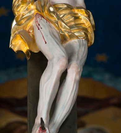Zdjęcie nr 14: Grupa Ukrzyżowania składa się z pełnoplastycznych, całopostaciowych figur ustawionych na belce tęczowej. Pośrodku zamocowano polichromowany na ciemnobrązowo krzyż z realistycznie rzeźbioną figurą Chrystusa o plastycznie opracowanym modelunku muskulatury ciała. Postać do krzyża przytwierdzono za pomocą trzech gwoździ. Zbawiciel ma głowę pochyloną i złożoną na prawym barku. Posiada szeroką, owalną twarz o sumarycznie opracowanych rysach, półprzymknięte oczy, lekko rozchylone usta i krótki, ciemnobrązowy zarost. Długie, ciemnobrązowe, falowane włosy opadają mu na plecy i na prawy bark. Na głowie znajduje się zielona, cierniowa korona, spod której ciekną stróżki krwi. Nieproporcjonalnie długie ramiona ułożone zostały w płytkim, U-kształtnym zwisie z zaznaczonymi ścięgnami i żyłami na przedramionach. Dłonie o zaciśniętych palcach przybito dwoma gwoździami, spod których ciekną strużki krwi. Klatkę piersiową ukształtowano z wyraziście zaznaczoną linią żeber i raną w boku ze strugami krwi. Na biodrach umiejscowiono krótkie, złocone perizonium z festonem przy prawym boku. Jego zwichrowane draperie zostały ułożone horyzontalnie. Chrystus ma zgięte w kolanach nogi z założoną prawą stopą na lewą przybite jednym gwoździem. W górnej partii pionowej belki krzyża zawieszono biały titulus w formie banderoli z napisem: "INRI". 
U stóp krzyża, po lewej stronie, na niskiej, prostopadłościennej podstawie, ustawiono rzeźbę klęczącej św. Marii Magdaleny. Święta jest zwrócona profilem w prawo. Jej owalną twarz charakteryzują sumarycznie opracowane rysy. Ma długie brązowe, lekko falowane włosy opadające na plecy. Ubrana jest w długą, srebrzystobłękitną, drobno fałdowaną suknię związaną srebrnym paskiem. Oburącz ujmuje pionową belkę krzyża. Tuż obok niej, na lewo, na niskiej, prostopadłościennej podstawie ustawiono figurę stojącej frontalnie Matki Boskiej Bolesnej. Jej nieznacznie odchyloną głowę charakteryzuje owalna, szeroka twarz o sumarycznie opracowanych rysach i wzroku skierowanym ku górze. Maria ubrana jest w długą srebrzyścieczerwoną suknię przewiązaną złotym paskiem i osłaniający głowę, plecy i prawe ramię, drobnofałdowany srebrzysty płaszcz. Ręce trzyma złożone w geście modlitwy. Po prawej stronie krzyża na niskiej, prostopadłościennej podstawie zamocowano stojącą figurę św. Jana Ewangelisty, ujętego frontalnie w nieznacznym kontrapoście. Święty ma lekko odchyloną do tyłu, zwróconą w trzech czwartych w lewo głowę. Charakteryzuje ją owalna, szeroka twarz o sumarycznie opracowanych rysach. Na plecy opadają mu długie, falowane, brązowe włosy. Jan ubrany jest w długą, srebrzystozieloną tunikę i przerzucony przez prawe ramię i podtrzymywany lewym przedramieniem złocistoochrowy płaszcz. Ręce trzyma skrzyżowane na piersiach. Z tyłu titulusa widnieje napis: Malował Jan i Ludwik, bracia Stankiewicze 1913. Napis widoczny jest także na górnej płaszczyźnie z tyłu krzyża: Malował 1913. 
Belka tęczowa jest prostopadłościenna, o profilowanych dolnych narożach. Polichromowano ją w odcieniach zieleni, czerwieni i żółci. Na środku widnieje flankowany parą złoconych krzyżyków złocony z przodu napis: + OTO BARANEK BOŻY KTÓRY GŁADZI GRZECHY ŚWIATA +; z tyłu: RENOVATUM A[nno] D[omini] 1913 + 1953 + 1983 + 2013