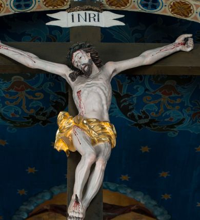 Zdjęcie nr 11: Grupa Ukrzyżowania składa się z pełnoplastycznych, całopostaciowych figur ustawionych na belce tęczowej. Pośrodku zamocowano polichromowany na ciemnobrązowo krzyż z realistycznie rzeźbioną figurą Chrystusa o plastycznie opracowanym modelunku muskulatury ciała. Postać do krzyża przytwierdzono za pomocą trzech gwoździ. Zbawiciel ma głowę pochyloną i złożoną na prawym barku. Posiada szeroką, owalną twarz o sumarycznie opracowanych rysach, półprzymknięte oczy, lekko rozchylone usta i krótki, ciemnobrązowy zarost. Długie, ciemnobrązowe, falowane włosy opadają mu na plecy i na prawy bark. Na głowie znajduje się zielona, cierniowa korona, spod której ciekną stróżki krwi. Nieproporcjonalnie długie ramiona ułożone zostały w płytkim, U-kształtnym zwisie z zaznaczonymi ścięgnami i żyłami na przedramionach. Dłonie o zaciśniętych palcach przybito dwoma gwoździami, spod których ciekną strużki krwi. Klatkę piersiową ukształtowano z wyraziście zaznaczoną linią żeber i raną w boku ze strugami krwi. Na biodrach umiejscowiono krótkie, złocone perizonium z festonem przy prawym boku. Jego zwichrowane draperie zostały ułożone horyzontalnie. Chrystus ma zgięte w kolanach nogi z założoną prawą stopą na lewą przybite jednym gwoździem. W górnej partii pionowej belki krzyża zawieszono biały titulus w formie banderoli z napisem: "INRI". 
U stóp krzyża, po lewej stronie, na niskiej, prostopadłościennej podstawie, ustawiono rzeźbę klęczącej św. Marii Magdaleny. Święta jest zwrócona profilem w prawo. Jej owalną twarz charakteryzują sumarycznie opracowane rysy. Ma długie brązowe, lekko falowane włosy opadające na plecy. Ubrana jest w długą, srebrzystobłękitną, drobno fałdowaną suknię związaną srebrnym paskiem. Oburącz ujmuje pionową belkę krzyża. Tuż obok niej, na lewo, na niskiej, prostopadłościennej podstawie ustawiono figurę stojącej frontalnie Matki Boskiej Bolesnej. Jej nieznacznie odchyloną głowę charakteryzuje owalna, szeroka twarz o sumarycznie opracowanych rysach i wzroku skierowanym ku górze. Maria ubrana jest w długą srebrzyścieczerwoną suknię przewiązaną złotym paskiem i osłaniający głowę, plecy i prawe ramię, drobnofałdowany srebrzysty płaszcz. Ręce trzyma złożone w geście modlitwy. Po prawej stronie krzyża na niskiej, prostopadłościennej podstawie zamocowano stojącą figurę św. Jana Ewangelisty, ujętego frontalnie w nieznacznym kontrapoście. Święty ma lekko odchyloną do tyłu, zwróconą w trzech czwartych w lewo głowę. Charakteryzuje ją owalna, szeroka twarz o sumarycznie opracowanych rysach. Na plecy opadają mu długie, falowane, brązowe włosy. Jan ubrany jest w długą, srebrzystozieloną tunikę i przerzucony przez prawe ramię i podtrzymywany lewym przedramieniem złocistoochrowy płaszcz. Ręce trzyma skrzyżowane na piersiach. Z tyłu titulusa widnieje napis: Malował Jan i Ludwik, bracia Stankiewicze 1913. Napis widoczny jest także na górnej płaszczyźnie z tyłu krzyża: Malował 1913. 
Belka tęczowa jest prostopadłościenna, o profilowanych dolnych narożach. Polichromowano ją w odcieniach zieleni, czerwieni i żółci. Na środku widnieje flankowany parą złoconych krzyżyków złocony z przodu napis: + OTO BARANEK BOŻY KTÓRY GŁADZI GRZECHY ŚWIATA +; z tyłu: RENOVATUM A[nno] D[omini] 1913 + 1953 + 1983 + 2013