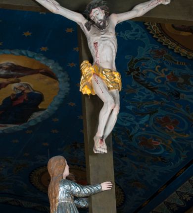 Zdjęcie nr 2: Grupa Ukrzyżowania składa się z pełnoplastycznych, całopostaciowych figur ustawionych na belce tęczowej. Pośrodku zamocowano polichromowany na ciemnobrązowo krzyż z realistycznie rzeźbioną figurą Chrystusa o plastycznie opracowanym modelunku muskulatury ciała. Postać do krzyża przytwierdzono za pomocą trzech gwoździ. Zbawiciel ma głowę pochyloną i złożoną na prawym barku. Posiada szeroką, owalną twarz o sumarycznie opracowanych rysach, półprzymknięte oczy, lekko rozchylone usta i krótki, ciemnobrązowy zarost. Długie, ciemnobrązowe, falowane włosy opadają mu na plecy i na prawy bark. Na głowie znajduje się zielona, cierniowa korona, spod której ciekną stróżki krwi. Nieproporcjonalnie długie ramiona ułożone zostały w płytkim, U-kształtnym zwisie z zaznaczonymi ścięgnami i żyłami na przedramionach. Dłonie o zaciśniętych palcach przybito dwoma gwoździami, spod których ciekną strużki krwi. Klatkę piersiową ukształtowano z wyraziście zaznaczoną linią żeber i raną w boku ze strugami krwi. Na biodrach umiejscowiono krótkie, złocone perizonium z festonem przy prawym boku. Jego zwichrowane draperie zostały ułożone horyzontalnie. Chrystus ma zgięte w kolanach nogi z założoną prawą stopą na lewą przybite jednym gwoździem. W górnej partii pionowej belki krzyża zawieszono biały titulus w formie banderoli z napisem: "INRI". 
U stóp krzyża, po lewej stronie, na niskiej, prostopadłościennej podstawie, ustawiono rzeźbę klęczącej św. Marii Magdaleny. Święta jest zwrócona profilem w prawo. Jej owalną twarz charakteryzują sumarycznie opracowane rysy. Ma długie brązowe, lekko falowane włosy opadające na plecy. Ubrana jest w długą, srebrzystobłękitną, drobno fałdowaną suknię związaną srebrnym paskiem. Oburącz ujmuje pionową belkę krzyża. Tuż obok niej, na lewo, na niskiej, prostopadłościennej podstawie ustawiono figurę stojącej frontalnie Matki Boskiej Bolesnej. Jej nieznacznie odchyloną głowę charakteryzuje owalna, szeroka twarz o sumarycznie opracowanych rysach i wzroku skierowanym ku górze. Maria ubrana jest w długą srebrzyścieczerwoną suknię przewiązaną złotym paskiem i osłaniający głowę, plecy i prawe ramię, drobnofałdowany srebrzysty płaszcz. Ręce trzyma złożone w geście modlitwy. Po prawej stronie krzyża na niskiej, prostopadłościennej podstawie zamocowano stojącą figurę św. Jana Ewangelisty, ujętego frontalnie w nieznacznym kontrapoście. Święty ma lekko odchyloną do tyłu, zwróconą w trzech czwartych w lewo głowę. Charakteryzuje ją owalna, szeroka twarz o sumarycznie opracowanych rysach. Na plecy opadają mu długie, falowane, brązowe włosy. Jan ubrany jest w długą, srebrzystozieloną tunikę i przerzucony przez prawe ramię i podtrzymywany lewym przedramieniem złocistoochrowy płaszcz. Ręce trzyma skrzyżowane na piersiach. Z tyłu titulusa widnieje napis: Malował Jan i Ludwik, bracia Stankiewicze 1913. Napis widoczny jest także na górnej płaszczyźnie z tyłu krzyża: Malował 1913. 
Belka tęczowa jest prostopadłościenna, o profilowanych dolnych narożach. Polichromowano ją w odcieniach zieleni, czerwieni i żółci. Na środku widnieje flankowany parą złoconych krzyżyków złocony z przodu napis: + OTO BARANEK BOŻY KTÓRY GŁADZI GRZECHY ŚWIATA +; z tyłu: RENOVATUM A[nno] D[omini] 1913 + 1953 + 1983 + 2013
