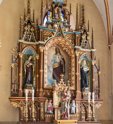 Zdjęcie nr 1: Jednokondygnacyjny i trójosiowy ołtarz ze zwieńczeniem uzyskał architektoniczną, bejcowaną i lakierowaną strukturę z partiami detalu i ornamentyką polichromowaną w kolorze czerwonym i niebieskim. Stipes ołtarzowy przyjął kształt prostopadłościanu z drewnianą mensą. Jego przód zdobią trzy pola z czworobocznymi płycinami ujętymi w złocone, profilowane ramki, rozdzielone kolumienkami z kapitelami korynckimi, ustawione na wysokich cokolikach z uskokiem. Środkową płaszczyznę dekoruje płaskorzeźbione i złocone przedstawienie Boskiego Serca w promienistej glorii z girlandą różaną. W dwóch pozostałych umieszczono płaskorzeźbione i złocone splecione litery Alfa i Omega oraz znak "Chi-Ro". Za ołtarzem rozpościera się wysoki cokół z parą niszy o konsolach zdobionych winną latoroślą. Na nim wspiera się predella nastawy dekorowana trójlistnymi arkadkami wspartymi na kolumienkach o złoconych korynckich kapitelach. W polach arkad umieszczono płaskorzeźbione i złocone przedstawienia stylizowanego kwiatu lilii. Na końcach predelli umieszczono ornament akantowy. Pośrodku predelli wkomponowano tabernakulum z tronem wystawienia. Tabernakulum uzyskało kształt prostopadłościennej szafki flankowanej parą złoconych kolumienek wspierających wyłamujący się ponad nimi gzyms. Dwuskrzydłowe drzwiczki zdobią płaskorzeźbione, złocone kłosy zbóż. Tabernakulum wieńczy złocony tron wystawienia w formie baldachimu wspartego na czterech kolumienkach, zamkniętego trzema ściankami z ośmioboku. Z kolei baldachim wieńczy ażurowa wimperga i para pinakli zdobione czołgankami i kwiatonami. Całość flankuje para prostopadłościennych cokołów zdobionych złoconym ornamentem, z dwoma figurami przyklękających na jednym kolanie aniołów z rękoma złożonymi w geście modlitewnym, ubranych w błękitno-różowe tuniki i dalmatyki. Każda z osi retabulum ujęta została parą kolumienek wspierających częściowo przerwane, wyłamujące się ponad nimi belkowanie. Gzyms w części środkowej uzyskał formę wimpergi. Retabulum po bokach zostało ujęte parą ażurowych uszaków z pojedynczymi kolumienkami o skręconych trzonach, zwieńczonych pojedynczymi pinaklami. Część środkowa zwieńczenia została ustawiona na cokole o zryzalitowanych bokach zwieńczonych parą pinakli. W szczycie usytuowano wsparty na parze kolumienek ażurowy baldachim z alternacji wimperg i pinakli, z wysokim, ażurowym pinaklem pośrodku. Nad osiami bocznymi retabulum usytuowano formy pary niewielkich kapliczek, których kolumienki wspierają wieńczące je duże, ażurowe pinakle oraz wimpergi flankowane parami mniejszych pinakli. Kapliczki ujęte są parami ażurowych wimperg u dołu oraz parami wysokich pinakli po bokach. Pinakle i wimpergi całej nastawy zdobione są złoconymi czołgankami i kwiatonami. Pole osi środkowej retabulum uzyskało formę płytkiej wnęki w kształcie stojącego prostokąta zamkniętego łukiem półkolistym z arkaturą. Ujmuje go profilowana, złocona rama zdobiona złoconą wicią winnej latorośli na ciemnoczerwonym tle. W środku umieszczono obraz Najświętsze Serce Pana Jezusa. Pola osi bocznych nastawy uzyskały formę trójbocznych niszy w kształcie stojących prostokątów zamkniętych łukiem ostrym o krawędzi zdobionej ażurowym ornamentem. Ścianki niszy pokrywa malowany, złocony astwerk na jasnozielonym tle, natomiast ich konchy błękit. Na ośmiobocznych postumentach zdobionych ornamentem ustawiono rzeźby świętych z podpisami. Z lewej strony znalazła się św. Teresa od Dzieciątka Jezus, a z prawej św. Małgorzata Alacoque. W zwieńczeniu, pośrodku, pod baldachimem umieszczono rzeźbę Chrystusa Dobrego Pasterza. Natomiast w kapliczkach po bokach parę figurek stojących aniołków ze złożonymi w geście modlitewnym rękoma.

