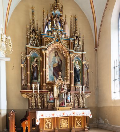 Zdjęcie nr 2: Jednokondygnacyjny i trójosiowy ołtarz ze zwieńczeniem uzyskał architektoniczną, bejcowaną i lakierowaną strukturę z partiami detalu i ornamentyką polichromowaną w kolorze czerwonym i niebieskim. Stipes ołtarzowy przyjął kształt prostopadłościanu z drewnianą mensą. Jego przód zdobią trzy pola z czworobocznymi płycinami ujętymi w złocone, profilowane ramki, rozdzielone kolumienkami z kapitelami korynckimi, ustawione na wysokich cokolikach z uskokiem. Środkową płaszczyznę dekoruje płaskorzeźbione i złocone przedstawienie Boskiego Serca w promienistej glorii z girlandą różaną. W dwóch pozostałych umieszczono płaskorzeźbione i złocone splecione litery Alfa i Omega oraz znak "Chi-Ro". Za ołtarzem rozpościera się wysoki cokół z parą niszy o konsolach zdobionych winną latoroślą. Na nim wspiera się predella nastawy dekorowana trójlistnymi arkadkami wspartymi na kolumienkach o złoconych korynckich kapitelach. W polach arkad umieszczono płaskorzeźbione i złocone przedstawienia stylizowanego kwiatu lilii. Na końcach predelli umieszczono ornament akantowy. Pośrodku predelli wkomponowano tabernakulum z tronem wystawienia. Tabernakulum uzyskało kształt prostopadłościennej szafki flankowanej parą złoconych kolumienek wspierających wyłamujący się ponad nimi gzyms. Dwuskrzydłowe drzwiczki zdobią płaskorzeźbione, złocone kłosy zbóż. Tabernakulum wieńczy złocony tron wystawienia w formie baldachimu wspartego na czterech kolumienkach, zamkniętego trzema ściankami z ośmioboku. Z kolei baldachim wieńczy ażurowa wimperga i para pinakli zdobione czołgankami i kwiatonami. Całość flankuje para prostopadłościennych cokołów zdobionych złoconym ornamentem, z dwoma figurami przyklękających na jednym kolanie aniołów z rękoma złożonymi w geście modlitewnym, ubranych w błękitno-różowe tuniki i dalmatyki. Każda z osi retabulum ujęta została parą kolumienek wspierających częściowo przerwane, wyłamujące się ponad nimi belkowanie. Gzyms w części środkowej uzyskał formę wimpergi. Retabulum po bokach zostało ujęte parą ażurowych uszaków z pojedynczymi kolumienkami o skręconych trzonach, zwieńczonych pojedynczymi pinaklami. Część środkowa zwieńczenia została ustawiona na cokole o zryzalitowanych bokach zwieńczonych parą pinakli. W szczycie usytuowano wsparty na parze kolumienek ażurowy baldachim z alternacji wimperg i pinakli, z wysokim, ażurowym pinaklem pośrodku. Nad osiami bocznymi retabulum usytuowano formy pary niewielkich kapliczek, których kolumienki wspierają wieńczące je duże, ażurowe pinakle oraz wimpergi flankowane parami mniejszych pinakli. Kapliczki ujęte są parami ażurowych wimperg u dołu oraz parami wysokich pinakli po bokach. Pinakle i wimpergi całej nastawy zdobione są złoconymi czołgankami i kwiatonami. Pole osi środkowej retabulum uzyskało formę płytkiej wnęki w kształcie stojącego prostokąta zamkniętego łukiem półkolistym z arkaturą. Ujmuje go profilowana, złocona rama zdobiona złoconą wicią winnej latorośli na ciemnoczerwonym tle. W środku umieszczono obraz Najświętsze Serce Pana Jezusa. Pola osi bocznych nastawy uzyskały formę trójbocznych niszy w kształcie stojących prostokątów zamkniętych łukiem ostrym o krawędzi zdobionej ażurowym ornamentem. Ścianki niszy pokrywa malowany, złocony astwerk na jasnozielonym tle, natomiast ich konchy błękit. Na ośmiobocznych postumentach zdobionych ornamentem ustawiono rzeźby świętych z podpisami. Z lewej strony znalazła się św. Teresa od Dzieciątka Jezus, a z prawej św. Małgorzata Alacoque. W zwieńczeniu, pośrodku, pod baldachimem umieszczono rzeźbę Chrystusa Dobrego Pasterza. Natomiast w kapliczkach po bokach parę figurek stojących aniołków ze złożonymi w geście modlitewnym rękoma.

