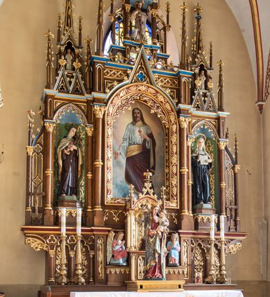 Zdjęcie nr 2: Obraz z przedstawieniem Najświętszego Serca Pana Jezusa uzyskał kształt stojącego prostokąta zamkniętego łukiem półkolistym. Ujmuje go profilowana, złocona rama zdobiona gałązkami różanymi. Jezus został ukazany frontalnie w całej postaci w lekkim kontrapoście, z lewą nogą nieznacznie odsuniętą w bok. Głowę ma lekko przechyloną w prawo. Zbawiciel prawą rękę ma opuszczoną wzdłuż tułowia z gestem ukazującym ranę na dłoni, z kolei lewą wskazuje na płonące serce na piersi. Charakteryzuje go pociągła twarz o jasnej karnacji i łagodnych rysach, z długim, prostym nosem i krótkim, brązowym zaroście. Głowę okalają długie, jasno brązowe włosy z przedziałkiem pośrodku spływające na ramiona. Ubrany jest w białą, przewiązaną w pasie tunikę z długimi, szerokimi rękawami, odsłaniającą fragment lewej stopy. Tunikę przy szyi wykańcza złociste obramienie. Jezus na tunice ma nałożony ciemnoczerwony płaszcz z jasnożółtą podszewką, obszyty wąskim, złotym galonem, przerzucony przez lewą rękę, osłaniający biodra i nogi. Na jego piersi jaśnieje przebite i płonące serce otoczone koroną cierniową. W tle widoczne są obłoki - w dolnej partii jasnobłękitne, wyżej rozświetlone.