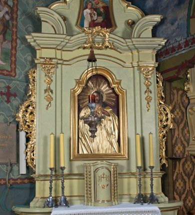 Zdjęcie nr 4: Obraz z wizerunkiem Najświętszego Serca Jezusa jest w kształcie stojącego prostokąta, od góry zamkniętego łukiem półkolistym, z górnymi narożnikami zaakcentowanymi przez wklęsłe łuki odcinkowe. Jezus został ukazany w półpostaci, niemal na wprost, głowę lekko przechyla w prawo. Pociągłą twarz charakteryzują delikatne rysy, długi prosty nos i krótki, ciemnobrązowy zarost. Długie, ciemnobrązowe, z przedziałkiem pośrodku, skręcone na końcach włosy opadają na ramiona. Dalsze elementy przedstawienia wydobyte są w obrębie partii złoconej sukienki: sylwetkę Jezusa osłania przewiązana pośrodku w pasie suknia o głęboko rozchylonym dekolcie; na ramiona narzucony jest płaszcz; dłońmi z ranami po gwoździach Jezus wskazuje na gorejące serce; jego głowę otacza nimb z wieńcem promieni. Tło jest jednolite, ciemnobordowe. Obraz ujęty jest profilowaną, złoconą ramą.