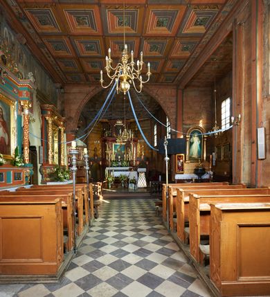 Zdjęcie nr 57: Kościół orientowany, pierwotnie na rzucie krzyża łacińskiego, jednonawowy, z kaplicami bocznymi od północy i południa, z prezbiterium prostokątnym zamkniętym wielobocznie. Obecnie w miejscu drewnianej kaplicy północnej murowana kaplica grobowa, a przy prezbiterium murowana prostokątna zakrystia i skarbczyk (1622 r.); nawa przedłużona ku zachodowi (1878-1879). Przy zachodniej części nawy, od południa, prostokątna drewniana kruchta. Wewnątrz stropy, w kaplicy północnej sklepienie krzyżowe z lunetami, w zakrystii i skarbczyku kolebkowe z lunetami. Zewnątrz zaszalowany. Dach nad nawą i prezbiterium jednokalenicowy, dwuspadowy o załamanych połaciach. Na kalenicy baniasta wieżyczka na sygnaturkę. Odrzwia zachodnie z nadprożem ozdobionym trójliściem z łukiem w ośli grzbiet oraz fryzem arkadkowym; odrzwia z prezbiterium do zakrystii z nadprożem ozdobionym łukiem w ośli grzbiet.