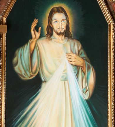 Zdjęcie nr 1: Obraz w kształcie stojącego prostokąta zamkniętego pięcioma bokami z ośmioboku. W centrum postać Chrystusa zwróconego en face, prawą ręką dotyka rany na piersi, lewą unosi w geście błogosławieństwa. Ubrany w białą szatę o szerokich rękawach, przepasaną w talii. Z boku wydobywają się wiązki promieni. Postać stoi na podłodze. Tło abstrakcyjne. U spodu obrazu aplikowana banderola z napisem: JEZU UFAM TOBIE.