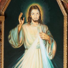 Zdjęcie nr 1: Obraz w kształcie stojącego prostokąta zamkniętego pięcioma bokami z ośmioboku. W centrum postać Chrystusa zwróconego en face, prawą ręką dotyka rany na piersi, lewą unosi w geście błogosławieństwa. Ubrany w białą szatę o szerokich rękawach, przepasaną w talii. Z boku wydobywają się wiązki promieni. Postać stoi na podłodze. Tło abstrakcyjne. U spodu obrazu aplikowana banderola z napisem: JEZU UFAM TOBIE.