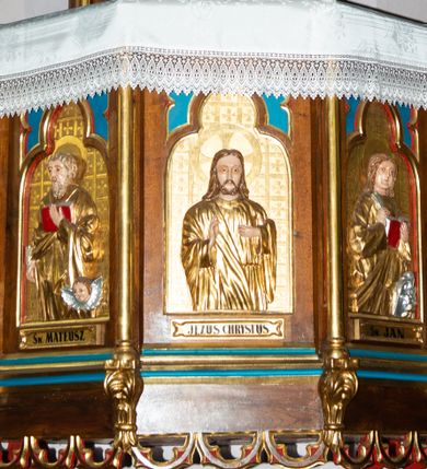 Zdjęcie nr 1: Ambona przyścienna, wisząca, z wejściem od strony zakrystii. Kosz na planie ośmioboku; ścianki wydzielone wąskimi kolumienkami, stojącymi na wspornikach; w polach płyciny w formie stojącego prostokąta, zamkniętego łukiem trójlistnym z maswerkami w przyłuczach, wewnątrz płaskorzeźbione półpostacie: św. Łukasza, św. Mateusza, Jezusa Chrystusa, św. Jana i św. Marka. Ewangeliści ukazani są frontalnie w trzech czwartych, z księgami oraz z piórami do pisania w dłoniach. Jezus z dłońmi ułożonymi w geście błogosławieństwa. Ubrani są w złote suknie i płaszcze. Wokół głów mają złote i koliste nimby; Jezus nimb krzyżowy. Ewangelistom towarzyszą ich symbole: św. Łukaszowi – byk, św. Mateuszowi – uskrzydlona główka aniołka, św. Janowi – orzeł, a św. Markowi – lew. Pod każdą płyciną znajdują się banderole z podpisami, kolejno od lewej: „Św(ięty) ŁUKASZ”, „Św(ięty) MATEUSZ”, „JEZUS CHRYSTUS”, „Św(ięty) JAN”, „Św(ięty) MAREK”. Tła płycin zapełnia kratownica, której kwadratowe pola wypełniają greckie krzyżyki. Dolna krawędź kosza zdobiona jest neogotyckim fartuchem; parapet ma formę gzymsu. 

Zaplecek wąski, ujęty parą kolumienek; w polu drzwi o kształcie prostokąta stojącego dekorowane dwiema płycinami z maswerkami; nad nimi prostokątna płaskorzeźba z przedstawieniem płynącej łodzi z monogramem Chrystusa na tle rozpiętego żagla. 

Baldachim powtarzający plan kosza, w formie podstawy dla ośmiobocznej ażurowej wieżyczki z blendami w ściankach i łukami przyporowymi, dekorowanej żabkami. Krawędź baldachimu profilowana, dekorowana sterczynami w narożach i neogotyckimi fartuchami u spodu i u góry. W podniebiu rzeźbiona gołębica Ducha Świętego w glorii.  

Struktura w kolorze naturalnego drewna; płyciny polichromowane na niebiesko i czerwono; elementy architektoniczne, profile, kolumienki i ścianki kosza złocone.

