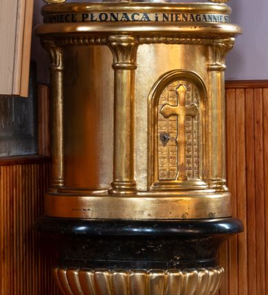 Zdjęcie nr 1: Chrzcielnica ustawiona na niskim cokole, na rzucie kwadratu; wsparta na półsferycznej stopie z cokolikiem, z datą wyrytą na płaszczu: „R(oku) P(ańskiego) 1869”. Trzon cylindryczny, lekko zwężający się ku górze, ozdobiony pionowymi żłobieniami. Czara w kształcie kielicha, w dolnej części puklowana, w górnej z wywiniętym silnie brzegiem. Pokrywa wysoka, w typie cylindrycznej aediculi ujętej kolumnami, podtrzymującymi wydatny gzyms. Na ściance frontowej drzwiczki w kształcie stojącego prostokąta, zamknięte łukiem półkolistym, ozdobione motywem kratownicy wypełnionej gwiazdkami i krzyżem o trójlistnie zakończonych ramionach. Na pokrywie grupa rzeźbiarska przedstawiająca Chrzest Chrystusa pod trzema wolutowymi kabłąkami spiętymi u góry koroną w typie zamkniętym. Nad gzymsem wtórny napis: „NA MILLENIUM 966-1966 WEŹMIJ ŚWIECĘ PŁONĄCĄ I NIENAGANNIE STRZEŻ CHRZTU SWOJEGO”. Pokrywa, kanelury oraz puklowanie złocone.
