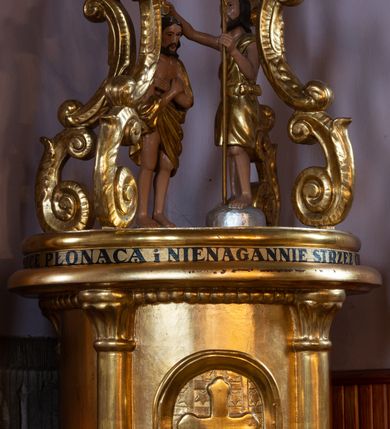 Zdjęcie nr 2: Chrzcielnica ustawiona na niskim cokole, na rzucie kwadratu; wsparta na półsferycznej stopie z cokolikiem, z datą wyrytą na płaszczu: „R(oku) P(ańskiego) 1869”. Trzon cylindryczny, lekko zwężający się ku górze, ozdobiony pionowymi żłobieniami. Czara w kształcie kielicha, w dolnej części puklowana, w górnej z wywiniętym silnie brzegiem. Pokrywa wysoka, w typie cylindrycznej aediculi ujętej kolumnami, podtrzymującymi wydatny gzyms. Na ściance frontowej drzwiczki w kształcie stojącego prostokąta, zamknięte łukiem półkolistym, ozdobione motywem kratownicy wypełnionej gwiazdkami i krzyżem o trójlistnie zakończonych ramionach. Na pokrywie grupa rzeźbiarska przedstawiająca Chrzest Chrystusa pod trzema wolutowymi kabłąkami spiętymi u góry koroną w typie zamkniętym. Nad gzymsem wtórny napis: „NA MILLENIUM 966-1966 WEŹMIJ ŚWIECĘ PŁONĄCĄ I NIENAGANNIE STRZEŻ CHRZTU SWOJEGO”. Pokrywa, kanelury oraz puklowanie złocone.
