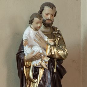 Zdjęcie nr 1: Rzeźba ustawiona na wysokim, profilowanym, prostopadłościennym cokole, w górnej części o ściętych narożach. Święty Józef został ukazany w postawie stojącej, w delikatnym kontrapoście, z prawą nogą ugiętą w kolanie, z Dzieciątkiem Jezus na prawym ręku, lewą ręką święty przyciska do piersi białą lilię, a dłonią podtrzymuje siedzącego Jezusa. Święty Józef ubrany w złotą, długą suknię z długimi rękawami oraz brązowy płaszcz. Jego twarz jest podłużna, o podkreślonych kościach policzkowych, z długim i wąskim nosem, małymi i drobnymi ustami, okolona średniej długości brodą, rozdzieloną na dwa pukle oraz średniej długości, ciemnobrązowymi włosami. Dzieciątko Jezus zwrócone trzy czwarte w lewo, w pozycji siedzącej, z rękami rozpostartymi szeroko na boki. Ubrane w długą, białą sukienkę, obwiedzioną na brzegach złotą lamówką. Twarz okrągła, z długim i szerokim nosem, z zarumienionymi policzkami; włosy jasnobrązowe, kędzierzawe, z wyraźnymi zakolami. Polichromia w odsłoniętych partiach ciała naturalistyczna. 
