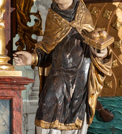 Zdjęcie nr 2: Pełnoplastyczne przedstawienie w kontrapoście ukazuje w całej postaci św. Mikołaja, biskupa. Święty ujęty został w trzech czwartych z głową widoczną w profilu. W nieznacznie odsuniętej od tułowia lewej ręce trzyma zamkniętą na dwie klamry złoconą księgę z leżącymi na niej obok siebie trzema złotymi kulami. Odsuniętą od tułowia ma także prawą rękę, w której pierwotnie trzymał pastorał, na co wskazuje układ palców dłoni. Mikołaj został ukazany jako dojrzały mężczyzna o jasnej karnacji. Ma pociągłą twarz bez zarostu z prostym nosem i wydatnymi ustami. Spod złotej mitry z fanonami opadającymi na plecy widać krótkie, brązowe włosy. Z przodu titulus mitry dekoruje płaskorzeźba mało czytelnej postaci, a circulus imitacja kameryzacji. Święty ubrany jest w mocno u spodu drapowaną albę, odsłaniającą wierzchołki ciemnych butów. Na albie nałożoną ma ciemną dalmatykę lamowaną u dołu złoconą frędzlą. Przykrywa ją złota kapa z dekoracyjną bortą zdobioną naśladownictwem kameryzacji i haftu, spiętą na piersi fibulą w kształcie rozety z kamieniem. 
