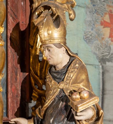 Zdjęcie nr 1: Pełnoplastyczne przedstawienie w kontrapoście ukazuje w całej postaci św. Mikołaja, biskupa. Święty ujęty został w trzech czwartych z głową widoczną w profilu. W nieznacznie odsuniętej od tułowia lewej ręce trzyma zamkniętą na dwie klamry złoconą księgę z leżącymi na niej obok siebie trzema złotymi kulami. Odsuniętą od tułowia ma także prawą rękę, w której pierwotnie trzymał pastorał, na co wskazuje układ palców dłoni. Mikołaj został ukazany jako dojrzały mężczyzna o jasnej karnacji. Ma pociągłą twarz bez zarostu z prostym nosem i wydatnymi ustami. Spod złotej mitry z fanonami opadającymi na plecy widać krótkie, brązowe włosy. Z przodu titulus mitry dekoruje płaskorzeźba mało czytelnej postaci, a circulus imitacja kameryzacji. Święty ubrany jest w mocno u spodu drapowaną albę, odsłaniającą wierzchołki ciemnych butów. Na albie nałożoną ma ciemną dalmatykę lamowaną u dołu złoconą frędzlą. Przykrywa ją złota kapa z dekoracyjną bortą zdobioną naśladownictwem kameryzacji i haftu, spiętą na piersi fibulą w kształcie rozety z kamieniem. 
