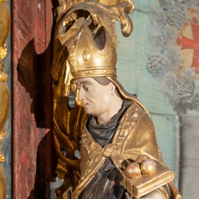 Zdjęcie nr 1: Pełnoplastyczne przedstawienie w kontrapoście ukazuje w całej postaci św. Mikołaja, biskupa. Święty ujęty został w trzech czwartych z głową widoczną w profilu. W nieznacznie odsuniętej od tułowia lewej ręce trzyma zamkniętą na dwie klamry złoconą księgę z leżącymi na niej obok siebie trzema złotymi kulami. Odsuniętą od tułowia ma także prawą rękę, w której pierwotnie trzymał pastorał, na co wskazuje układ palców dłoni. Mikołaj został ukazany jako dojrzały mężczyzna o jasnej karnacji. Ma pociągłą twarz bez zarostu z prostym nosem i wydatnymi ustami. Spod złotej mitry z fanonami opadającymi na plecy widać krótkie, brązowe włosy. Z przodu titulus mitry dekoruje płaskorzeźba mało czytelnej postaci, a circulus imitacja kameryzacji. Święty ubrany jest w mocno u spodu drapowaną albę, odsłaniającą wierzchołki ciemnych butów. Na albie nałożoną ma ciemną dalmatykę lamowaną u dołu złoconą frędzlą. Przykrywa ją złota kapa z dekoracyjną bortą zdobioną naśladownictwem kameryzacji i haftu, spiętą na piersi fibulą w kształcie rozety z kamieniem. 
