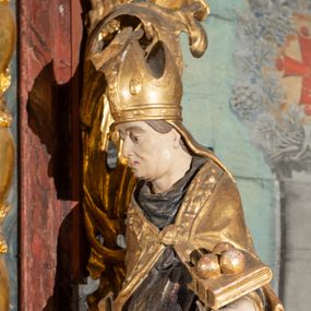 Zdjęcie nr 1: Pełnoplastyczne przedstawienie w kontrapoście ukazuje w całej postaci św. Mikołaja, biskupa. Święty ujęty został w trzech czwartych z głową widoczną w profilu. W nieznacznie odsuniętej od tułowia lewej ręce trzyma zamkniętą na dwie klamry złoconą księgę z leżącymi na niej obok siebie trzema złotymi kulami. Odsuniętą od tułowia ma także prawą rękę, w której pierwotnie trzymał pastorał, na co wskazuje układ palców dłoni. Mikołaj został ukazany jako dojrzały mężczyzna o jasnej karnacji. Ma pociągłą twarz bez zarostu z prostym nosem i wydatnymi ustami. Spod złotej mitry z fanonami opadającymi na plecy widać krótkie, brązowe włosy. Z przodu titulus mitry dekoruje płaskorzeźba mało czytelnej postaci, a circulus imitacja kameryzacji. Święty ubrany jest w mocno u spodu drapowaną albę, odsłaniającą wierzchołki ciemnych butów. Na albie nałożoną ma ciemną dalmatykę lamowaną u dołu złoconą frędzlą. Przykrywa ją złota kapa z dekoracyjną bortą zdobioną naśladownictwem kameryzacji i haftu, spiętą na piersi fibulą w kształcie rozety z kamieniem. 
