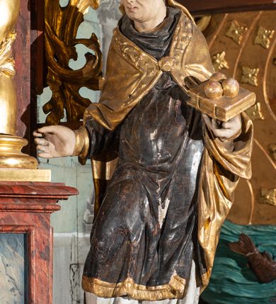 Zdjęcie nr 3: Pełnoplastyczne przedstawienie w kontrapoście ukazuje w całej postaci św. Mikołaja, biskupa. Święty ujęty został w trzech czwartych z głową widoczną w profilu. W nieznacznie odsuniętej od tułowia lewej ręce trzyma zamkniętą na dwie klamry złoconą księgę z leżącymi na niej obok siebie trzema złotymi kulami. Odsuniętą od tułowia ma także prawą rękę, w której pierwotnie trzymał pastorał, na co wskazuje układ palców dłoni. Mikołaj został ukazany jako dojrzały mężczyzna o jasnej karnacji. Ma pociągłą twarz bez zarostu z prostym nosem i wydatnymi ustami. Spod złotej mitry z fanonami opadającymi na plecy widać krótkie, brązowe włosy. Z przodu titulus mitry dekoruje płaskorzeźba mało czytelnej postaci, a circulus imitacja kameryzacji. Święty ubrany jest w mocno u spodu drapowaną albę, odsłaniającą wierzchołki ciemnych butów. Na albie nałożoną ma ciemną dalmatykę lamowaną u dołu złoconą frędzlą. Przykrywa ją złota kapa z dekoracyjną bortą zdobioną naśladownictwem kameryzacji i haftu, spiętą na piersi fibulą w kształcie rozety z kamieniem. 
