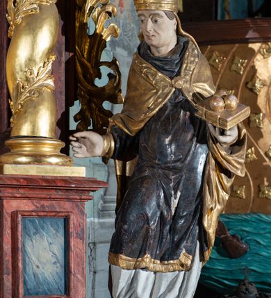 Zdjęcie nr 4: Pełnoplastyczne przedstawienie w kontrapoście ukazuje w całej postaci św. Mikołaja, biskupa. Święty ujęty został w trzech czwartych z głową widoczną w profilu. W nieznacznie odsuniętej od tułowia lewej ręce trzyma zamkniętą na dwie klamry złoconą księgę z leżącymi na niej obok siebie trzema złotymi kulami. Odsuniętą od tułowia ma także prawą rękę, w której pierwotnie trzymał pastorał, na co wskazuje układ palców dłoni. Mikołaj został ukazany jako dojrzały mężczyzna o jasnej karnacji. Ma pociągłą twarz bez zarostu z prostym nosem i wydatnymi ustami. Spod złotej mitry z fanonami opadającymi na plecy widać krótkie, brązowe włosy. Z przodu titulus mitry dekoruje płaskorzeźba mało czytelnej postaci, a circulus imitacja kameryzacji. Święty ubrany jest w mocno u spodu drapowaną albę, odsłaniającą wierzchołki ciemnych butów. Na albie nałożoną ma ciemną dalmatykę lamowaną u dołu złoconą frędzlą. Przykrywa ją złota kapa z dekoracyjną bortą zdobioną naśladownictwem kameryzacji i haftu, spiętą na piersi fibulą w kształcie rozety z kamieniem. 
