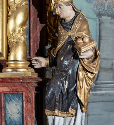 Zdjęcie nr 5: Pełnoplastyczne przedstawienie w kontrapoście ukazuje w całej postaci św. Mikołaja, biskupa. Święty ujęty został w trzech czwartych z głową widoczną w profilu. W nieznacznie odsuniętej od tułowia lewej ręce trzyma zamkniętą na dwie klamry złoconą księgę z leżącymi na niej obok siebie trzema złotymi kulami. Odsuniętą od tułowia ma także prawą rękę, w której pierwotnie trzymał pastorał, na co wskazuje układ palców dłoni. Mikołaj został ukazany jako dojrzały mężczyzna o jasnej karnacji. Ma pociągłą twarz bez zarostu z prostym nosem i wydatnymi ustami. Spod złotej mitry z fanonami opadającymi na plecy widać krótkie, brązowe włosy. Z przodu titulus mitry dekoruje płaskorzeźba mało czytelnej postaci, a circulus imitacja kameryzacji. Święty ubrany jest w mocno u spodu drapowaną albę, odsłaniającą wierzchołki ciemnych butów. Na albie nałożoną ma ciemną dalmatykę lamowaną u dołu złoconą frędzlą. Przykrywa ją złota kapa z dekoracyjną bortą zdobioną naśladownictwem kameryzacji i haftu, spiętą na piersi fibulą w kształcie rozety z kamieniem. 
