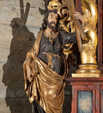 Zdjęcie nr 4: Pełnoplastyczne przedstawienie w kontrapoście ukazuje w całej postaci św. Andrzeja. Apostoł ujęty został w trzech czwartych. W uniesionej ku górze lewej ręce trzyma niewielki krzyż, określany jako krzyż św. Andrzeja. W prawej, opuszczonej wzdłuż tułowia i lekko zgiętej w łokciu, trzyma złoconą księgę zamkniętą na dwie klamry. Andrzej został ukazany jako dojrzały mężczyzna o jasnej karnacji. Ma pociągłą twarz okoloną brązowym zarostem oraz puklowane, brązowe włosy sięgające ramion. Twarz charakteryzują mocno otwarte oczy, wpatrzone w uniesiony krzyż, prosty nos oraz wydatne usta. Apostoł ubrany jest w mocno u spodu drapowaną, ciemną, luźną tunikę, wykończoną przy szyi złotą lamówką. Jest ona przewiązana w pasie złotą szarfą. Spod tuniki widać ciemne buty. Na ramionach ma narzucony złoty, mocno udrapowany płaszcz okrywający prawe ramię. Jego lewą połę przy prawym boku podtrzymuje ręką, w której trzyma księgę.   