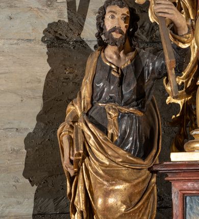 Zdjęcie nr 2: Pełnoplastyczne przedstawienie w kontrapoście ukazuje w całej postaci św. Andrzeja. Apostoł ujęty został w trzech czwartych. W uniesionej ku górze lewej ręce trzyma niewielki krzyż, określany jako krzyż św. Andrzeja. W prawej, opuszczonej wzdłuż tułowia i lekko zgiętej w łokciu, trzyma złoconą księgę zamkniętą na dwie klamry. Andrzej został ukazany jako dojrzały mężczyzna o jasnej karnacji. Ma pociągłą twarz okoloną brązowym zarostem oraz puklowane, brązowe włosy sięgające ramion. Twarz charakteryzują mocno otwarte oczy, wpatrzone w uniesiony krzyż, prosty nos oraz wydatne usta. Apostoł ubrany jest w mocno u spodu drapowaną, ciemną, luźną tunikę, wykończoną przy szyi złotą lamówką. Jest ona przewiązana w pasie złotą szarfą. Spod tuniki widać ciemne buty. Na ramionach ma narzucony złoty, mocno udrapowany płaszcz okrywający prawe ramię. Jego lewą połę przy prawym boku podtrzymuje ręką, w której trzyma księgę.   