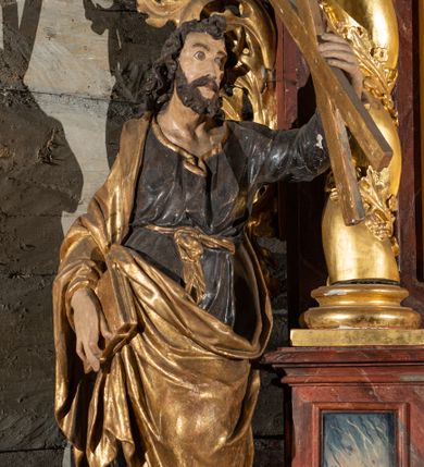 Zdjęcie nr 1: Pełnoplastyczne przedstawienie w kontrapoście ukazuje w całej postaci św. Andrzeja. Apostoł ujęty został w trzech czwartych. W uniesionej ku górze lewej ręce trzyma niewielki krzyż, określany jako krzyż św. Andrzeja. W prawej, opuszczonej wzdłuż tułowia i lekko zgiętej w łokciu, trzyma złoconą księgę zamkniętą na dwie klamry. Andrzej został ukazany jako dojrzały mężczyzna o jasnej karnacji. Ma pociągłą twarz okoloną brązowym zarostem oraz puklowane, brązowe włosy sięgające ramion. Twarz charakteryzują mocno otwarte oczy, wpatrzone w uniesiony krzyż, prosty nos oraz wydatne usta. Apostoł ubrany jest w mocno u spodu drapowaną, ciemną, luźną tunikę, wykończoną przy szyi złotą lamówką. Jest ona przewiązana w pasie złotą szarfą. Spod tuniki widać ciemne buty. Na ramionach ma narzucony złoty, mocno udrapowany płaszcz okrywający prawe ramię. Jego lewą połę przy prawym boku podtrzymuje ręką, w której trzyma księgę.   